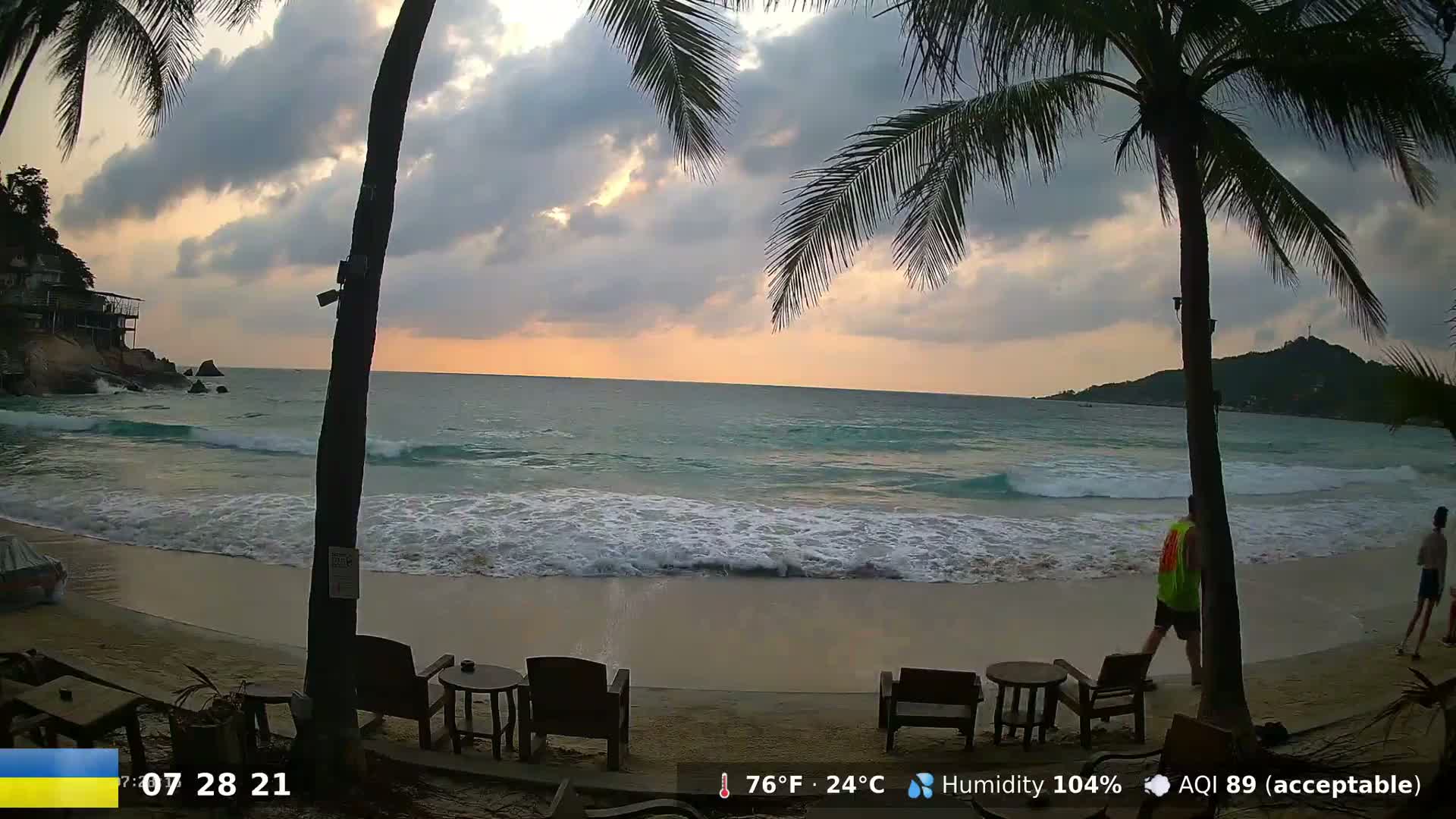 A sandy beach with gentle waves, palm trees, and outdoor seating at sunset under partly cloudy skies.