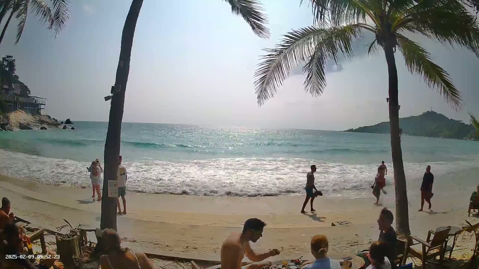 A sandy beach with turquoise water and gentle waves laps the shore under a sunny sky, where people relax and stroll near palm trees.
