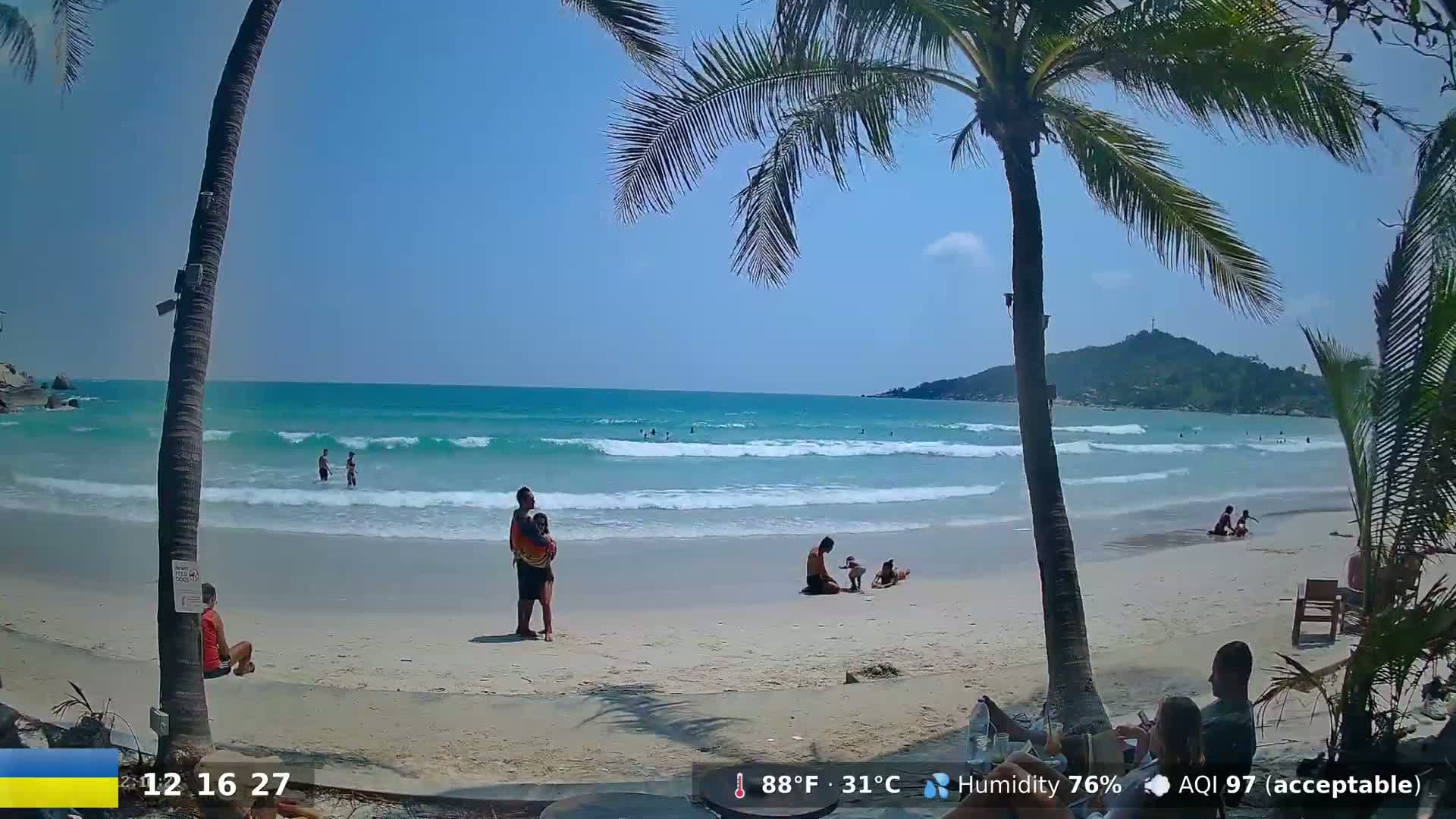 A tropical beach scene shows people relaxing on a white sand beach under a sunny sky with gentle waves.