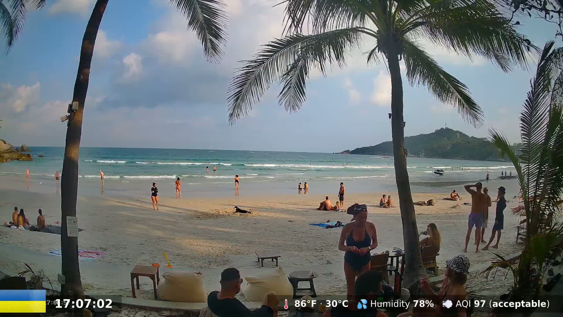 A sandy beach with palm trees and many people enjoying a partly cloudy day.