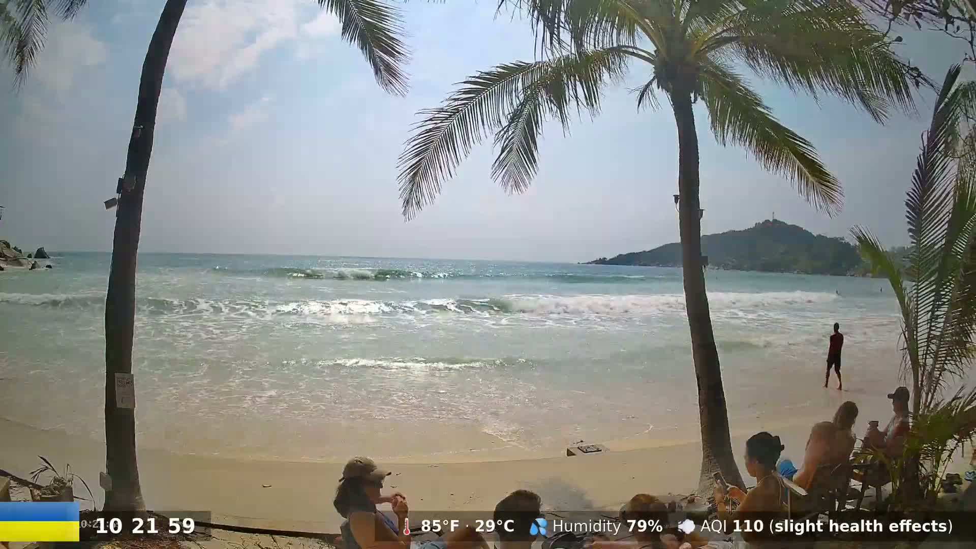 A sandy beach with gentle waves rolling in, palm trees framing the view, and several people relaxing on the shore under a partly sunny sky.