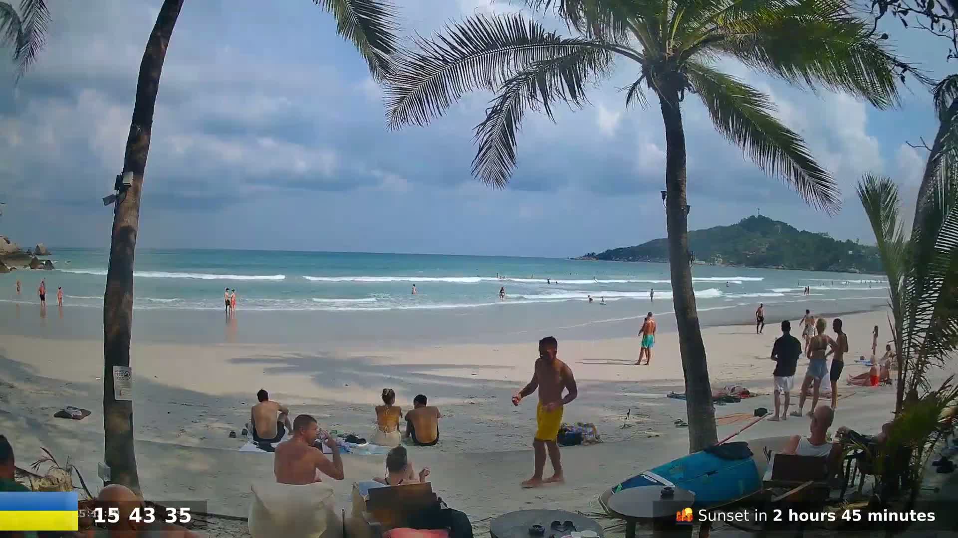 A sandy beach with a few people swimming and relaxing on a partly cloudy day, palm trees partially obscuring the view.