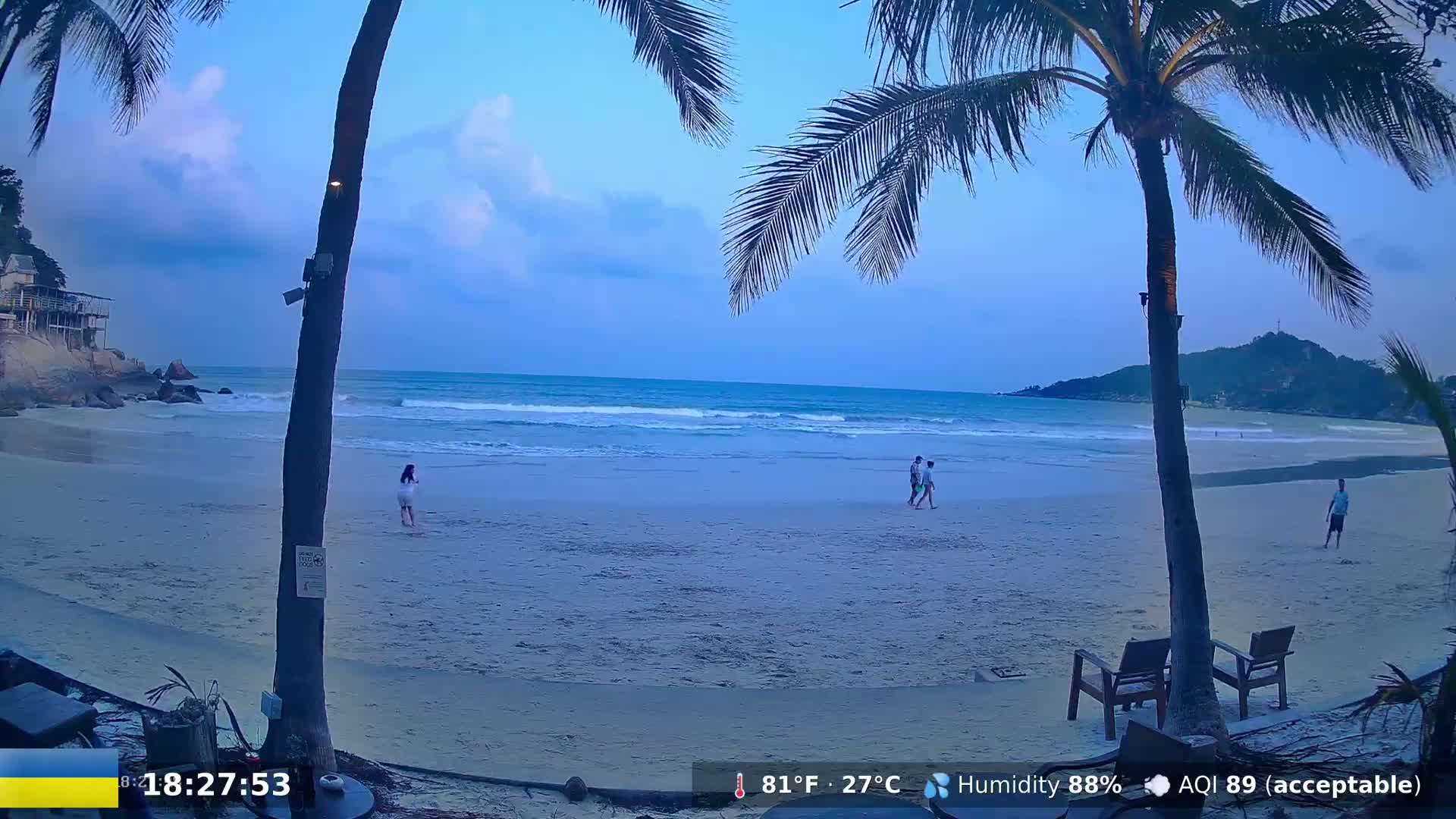 A sandy beach at dusk, under a mostly clear sky, with palm trees in the foreground and several people walking along the shore.
