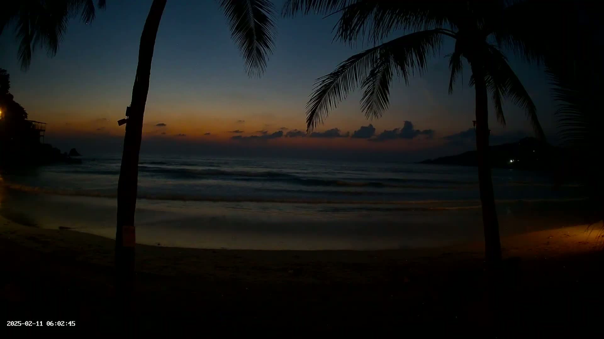 A dark beach at twilight shows gentle waves lapping the shore, framed by silhouetted palm trees under a dusky sky.