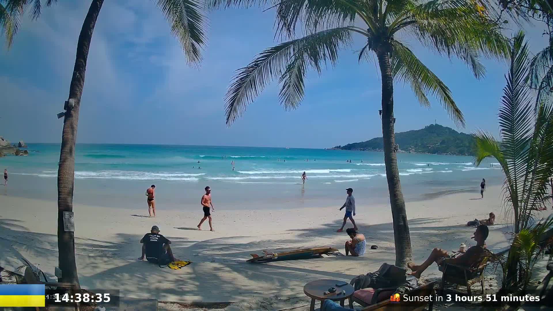 A sandy beach with palm trees, turquoise water with small waves, and several people swimming, sunbathing, and relaxing under a sunny sky.