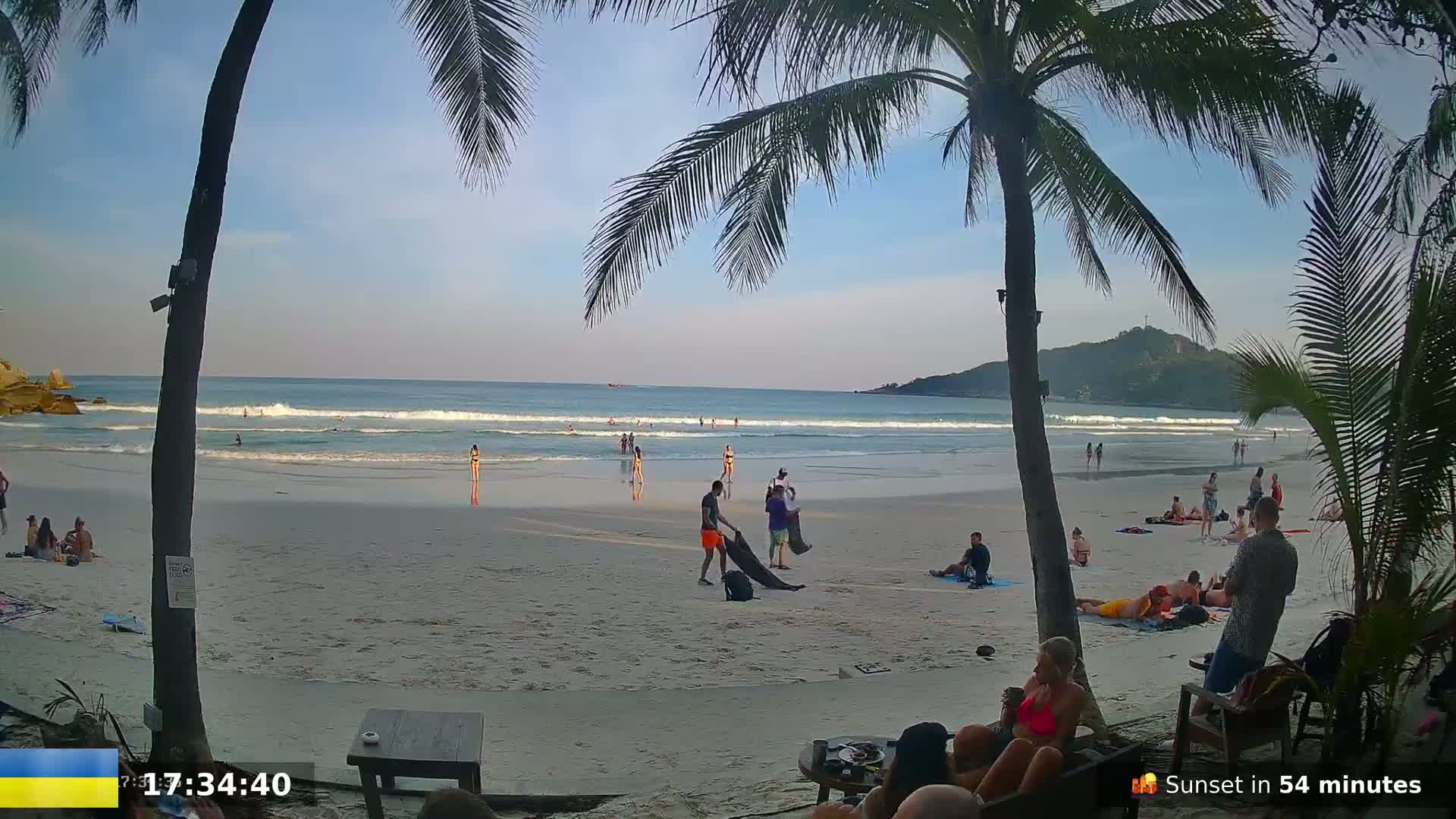A sandy beach with calm waves, a few people swimming and relaxing, palm trees framing the view under a mostly clear sky.
