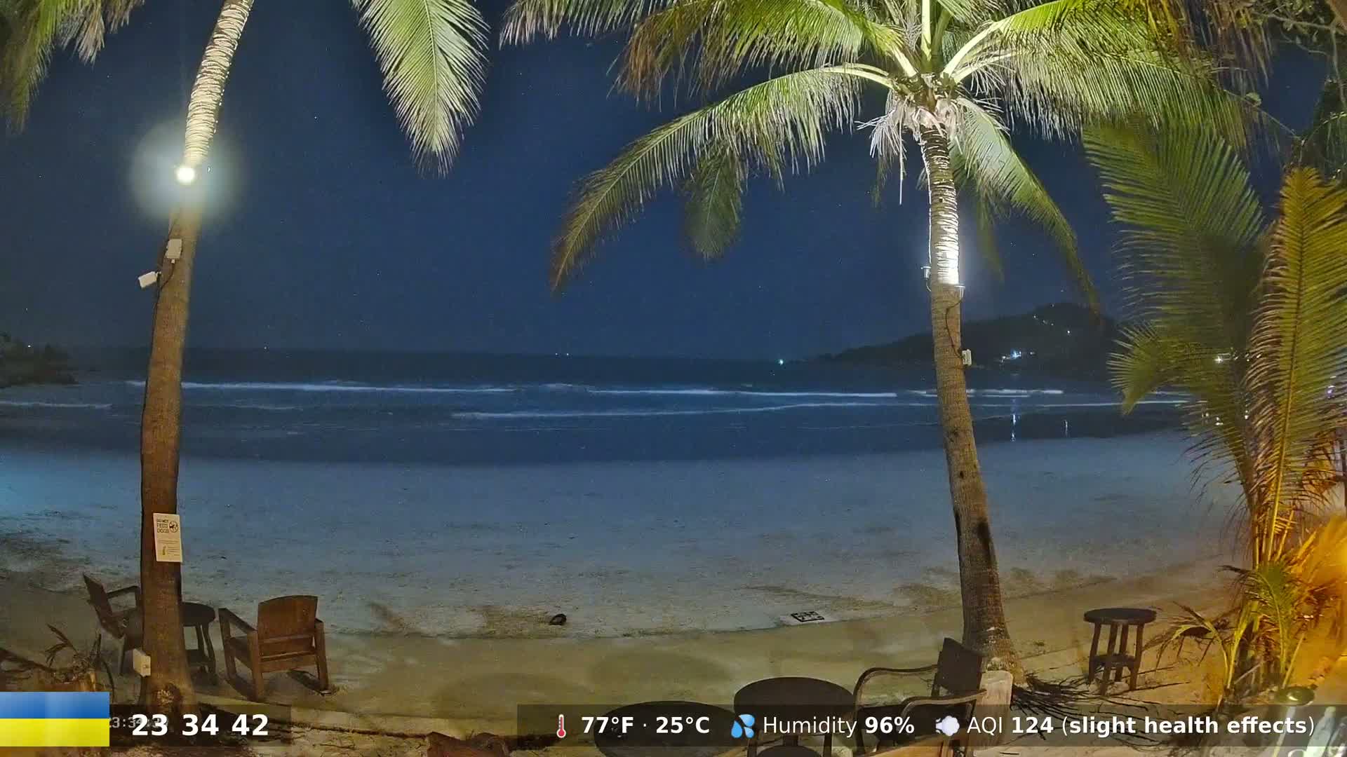 A nighttime beach scene shows a calm ocean under a dark, starry sky, with palm trees and outdoor furniture on the sand.