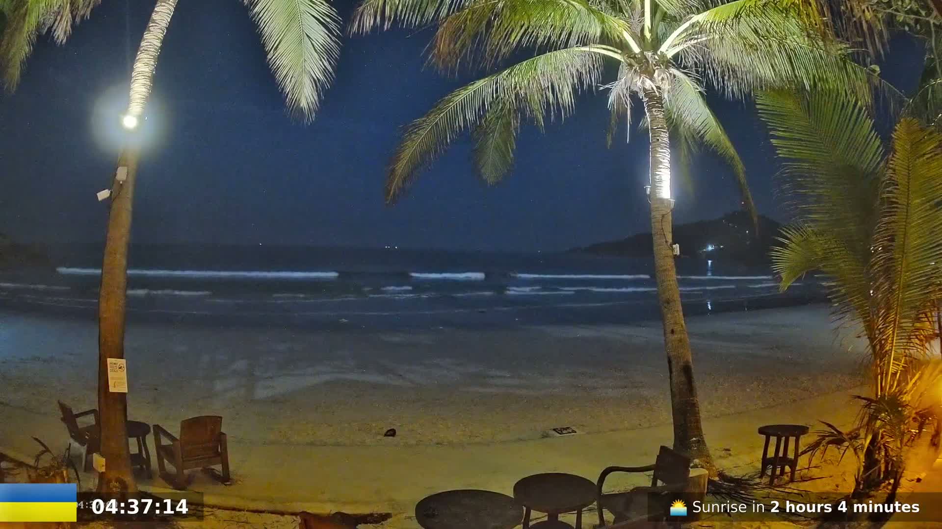 A nighttime beach scene shows gentle waves lapping the shore under a dark, starry sky, with palm trees and outdoor furniture visible in the foreground.