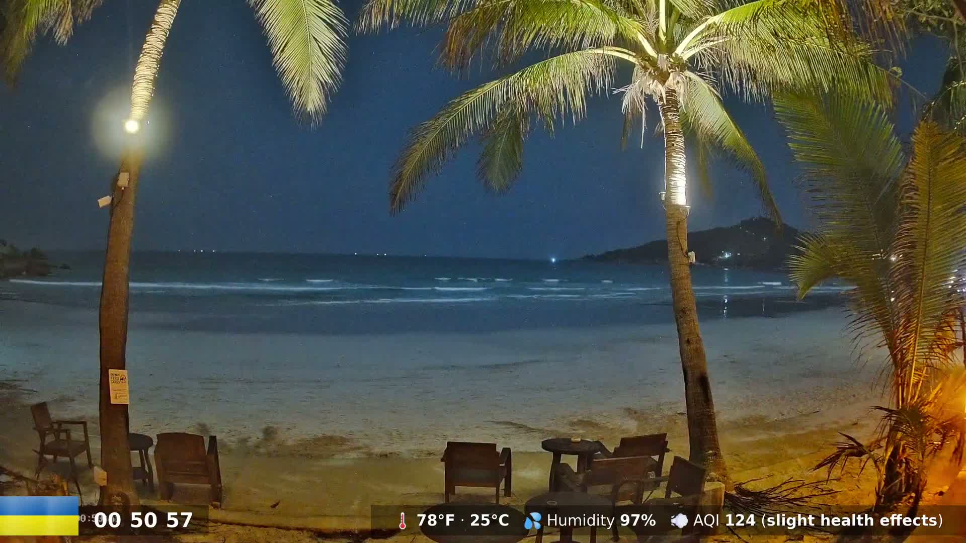 Under a dark, starry night sky, a tranquil beach scene shows palm trees illuminating a sandy shore with calm waves lapping at the edge, and a few outdoor tables and chairs situated on the beach.