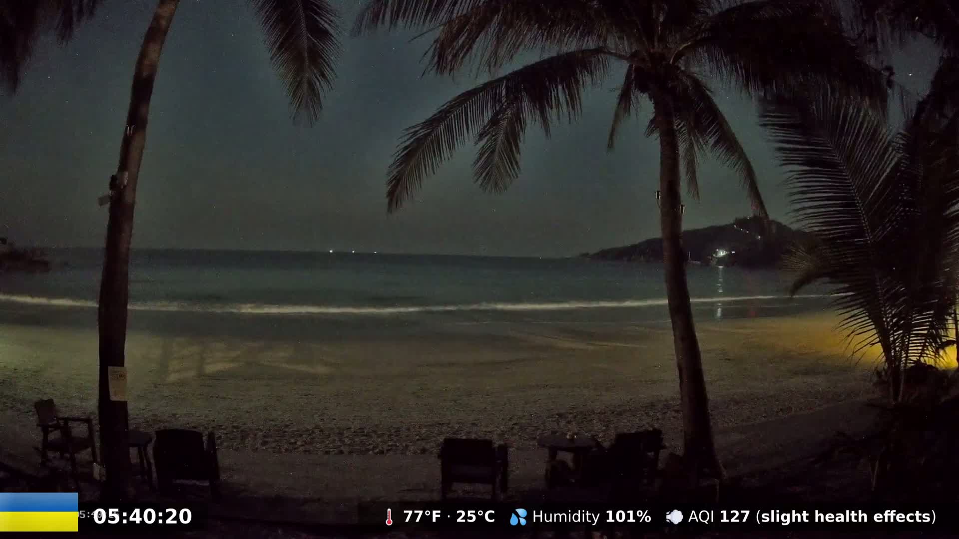 A dark, nighttime beach scene shows a calm ocean under a partly cloudy sky, viewed from between two palm trees with empty chairs on the sand.