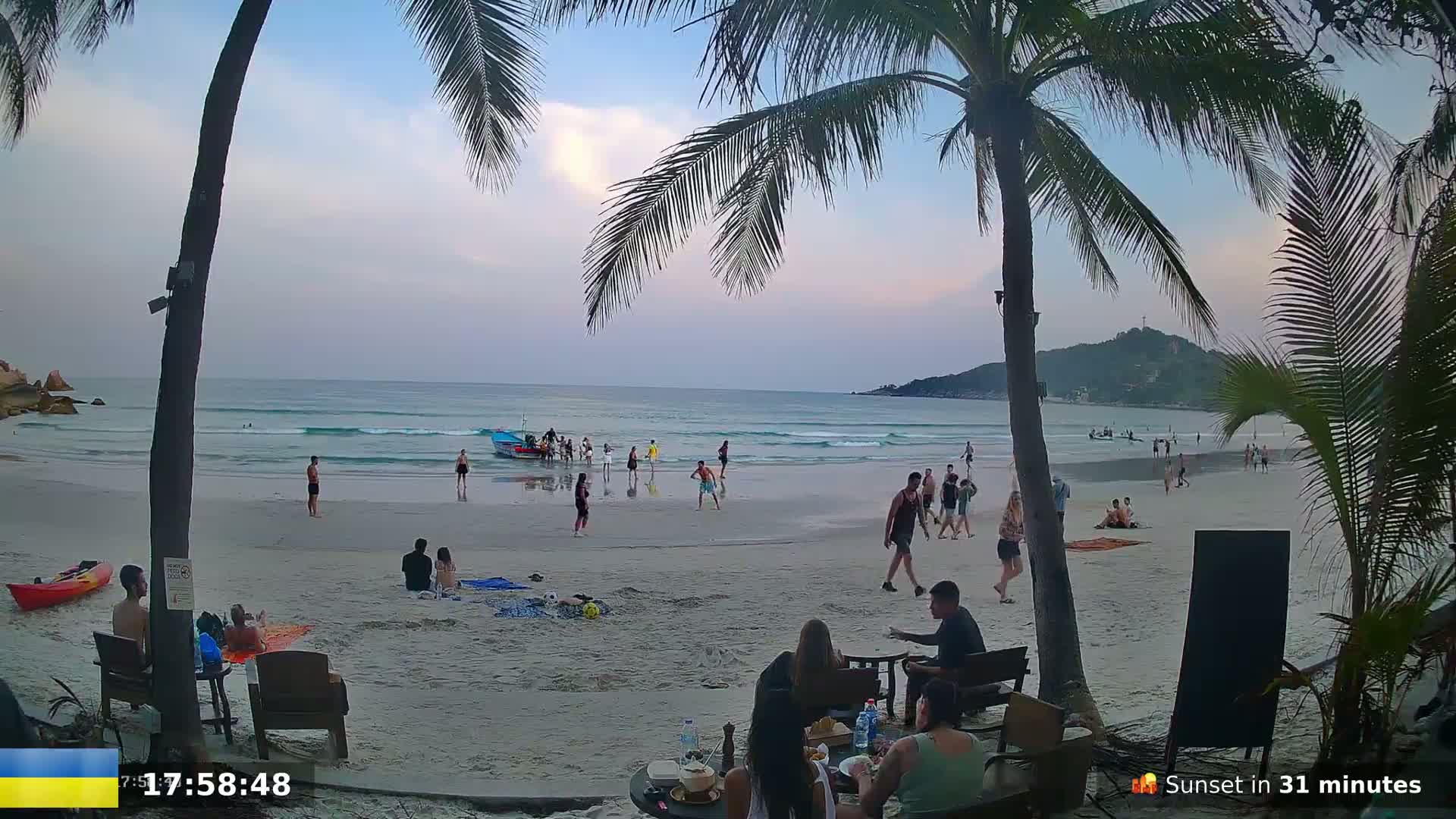 A sandy beach with people swimming, playing, and relaxing under a partly cloudy, dusky sky, as viewed from behind palm trees near an oceanside restaurant.