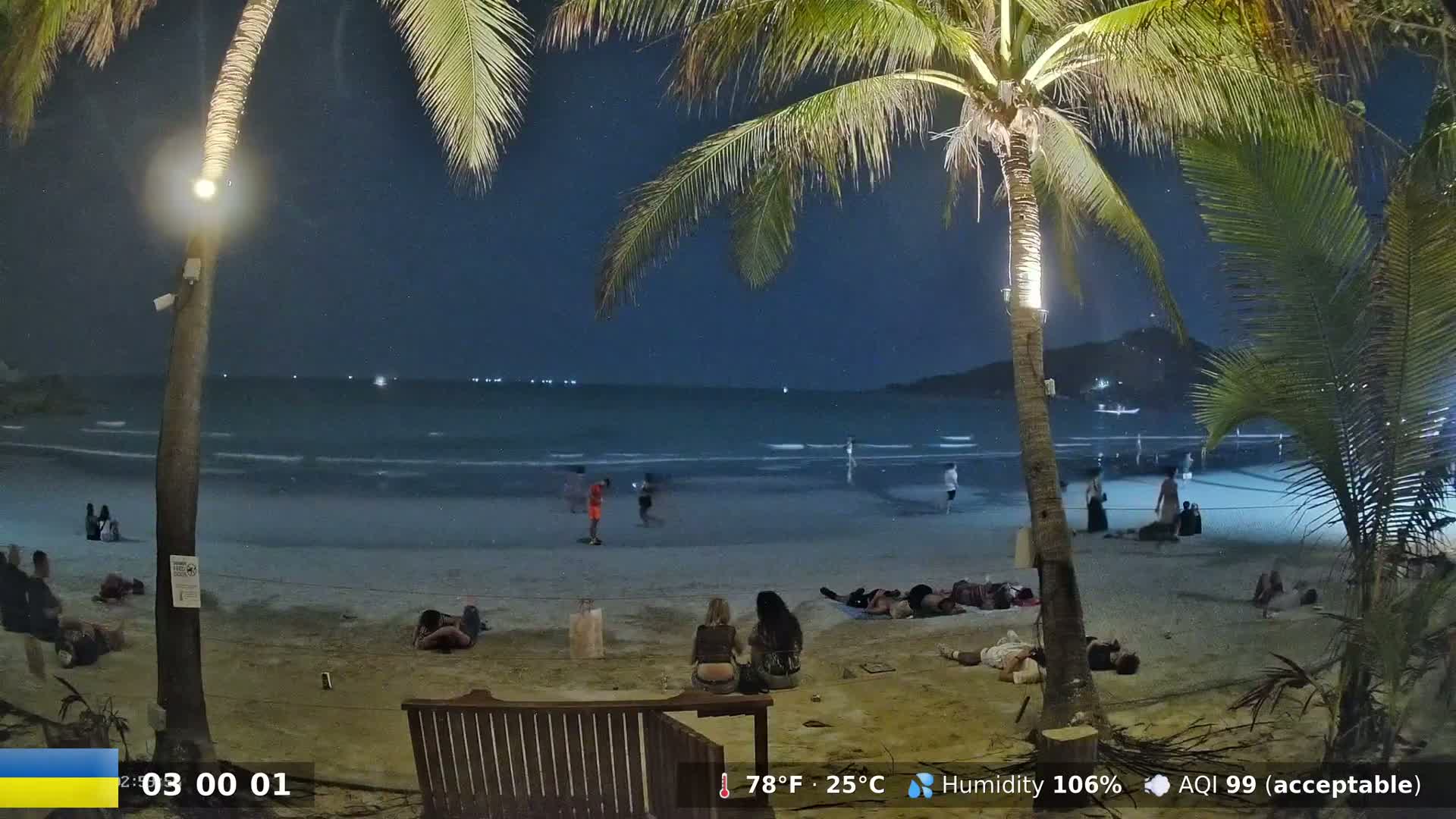 Under a dark, starry night sky, people relax and stroll along a sandy beach with calm ocean waves.