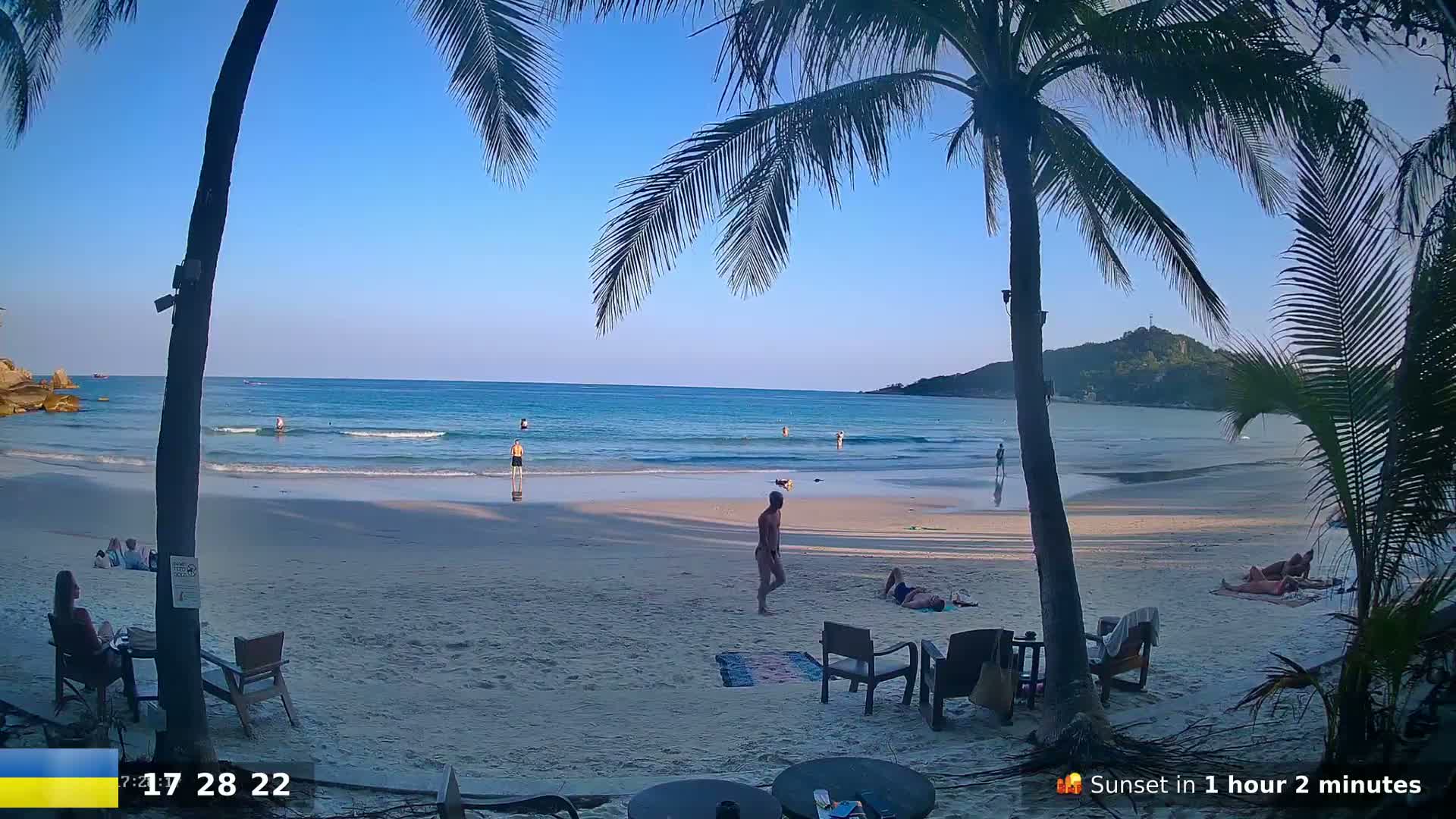 A sandy beach with calm ocean waves, several people swimming and relaxing, and palm trees framing the view under a clear blue sky.