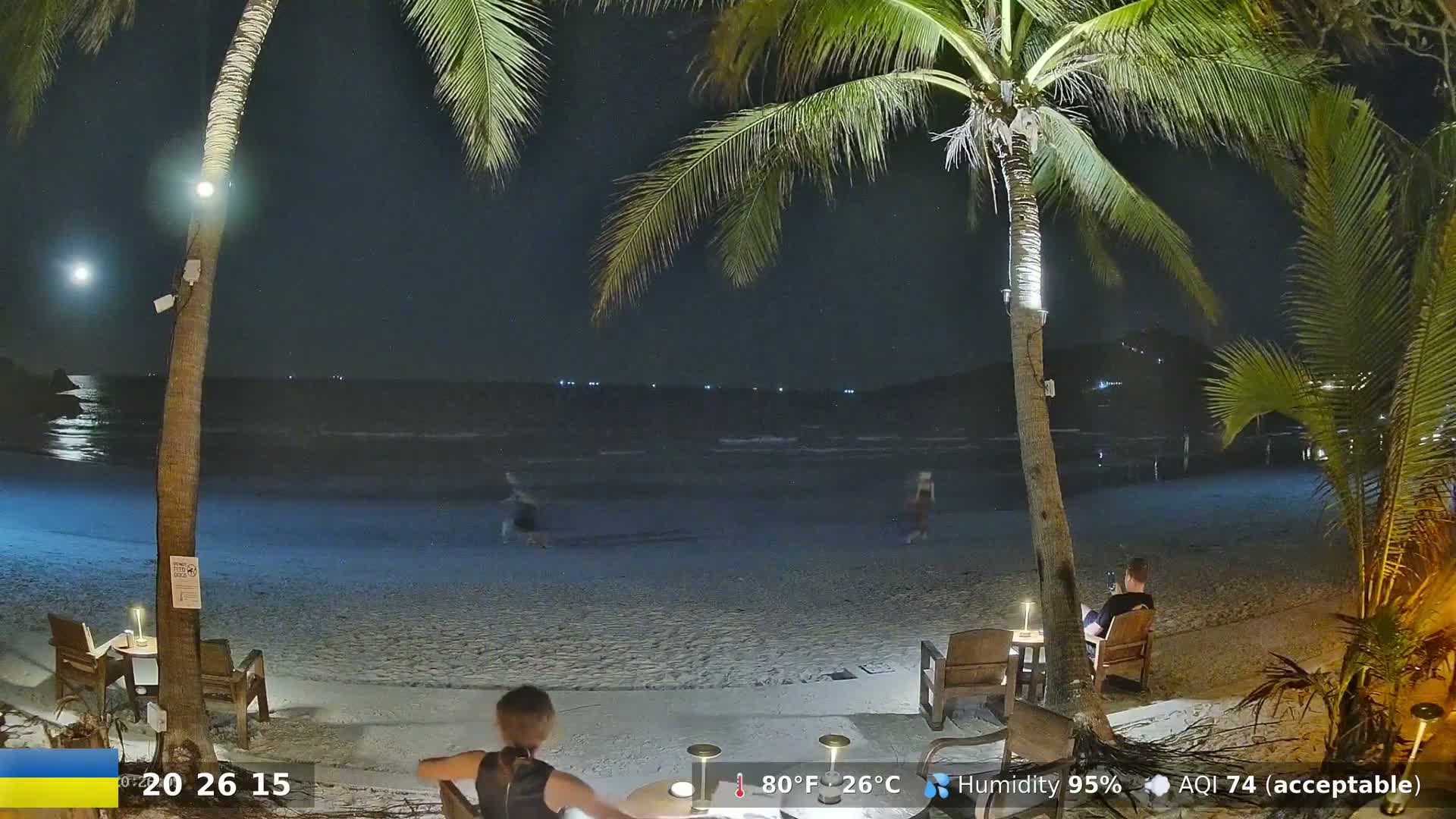 A nighttime beach scene shows a sandy shore, palm trees, a calm ocean, and several people sitting at tables under the palm trees.