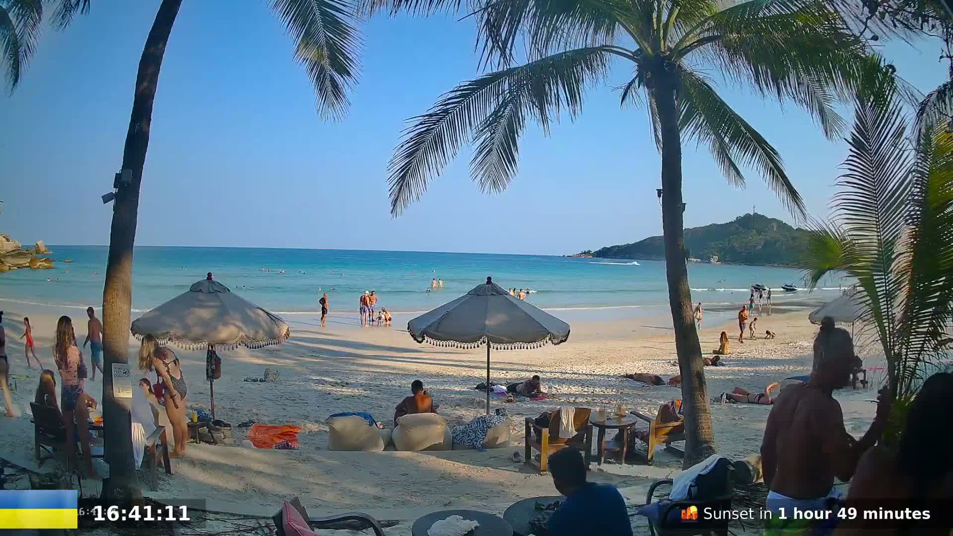 A sunny beach scene shows people relaxing under umbrellas and in the shallow water, with palm trees in the foreground and a small island visible in the distance.