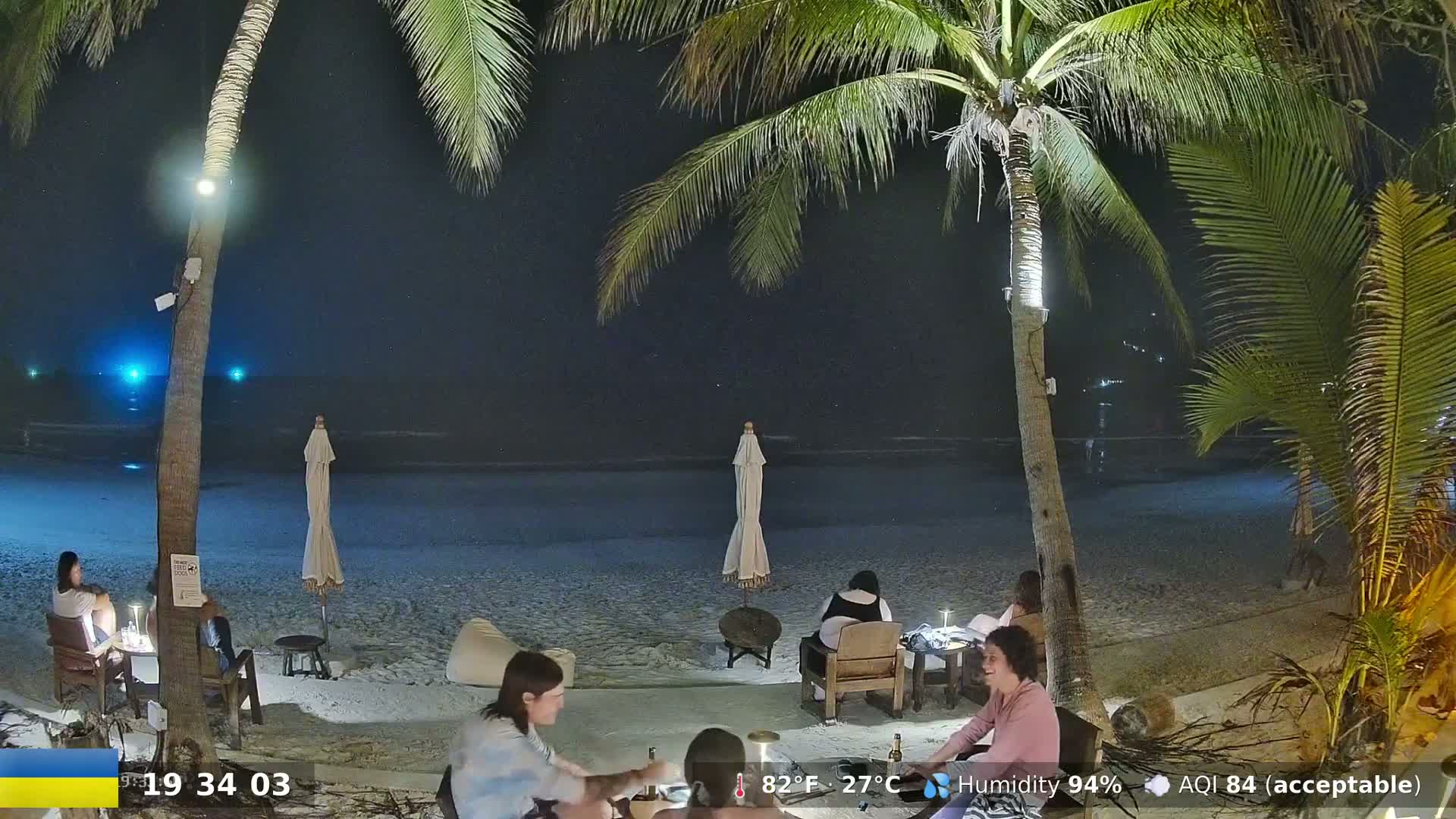 Under a clear night sky, several people sit at tables on a sandy beach illuminated by artificial lights, near palm trees and beach umbrellas.