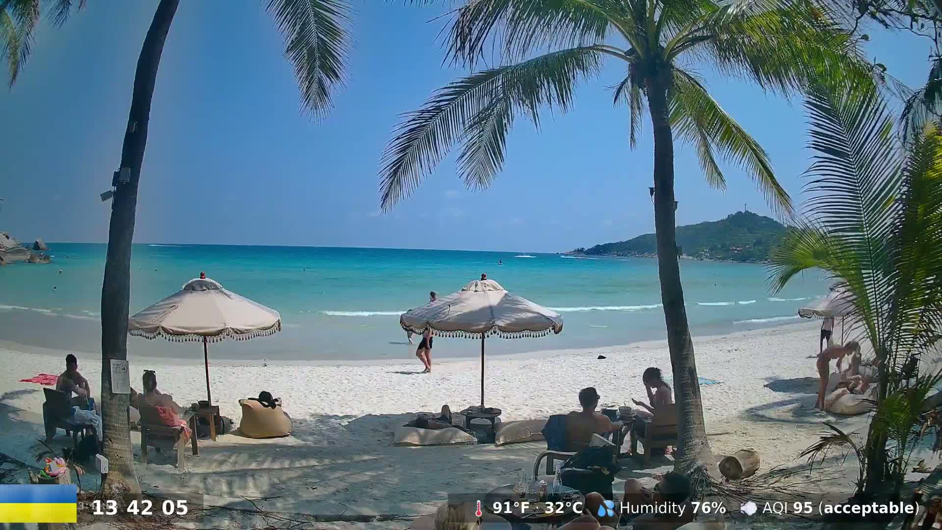 A sunny beach scene with turquoise water, palm trees, and several people relaxing under umbrellas on the white sand.