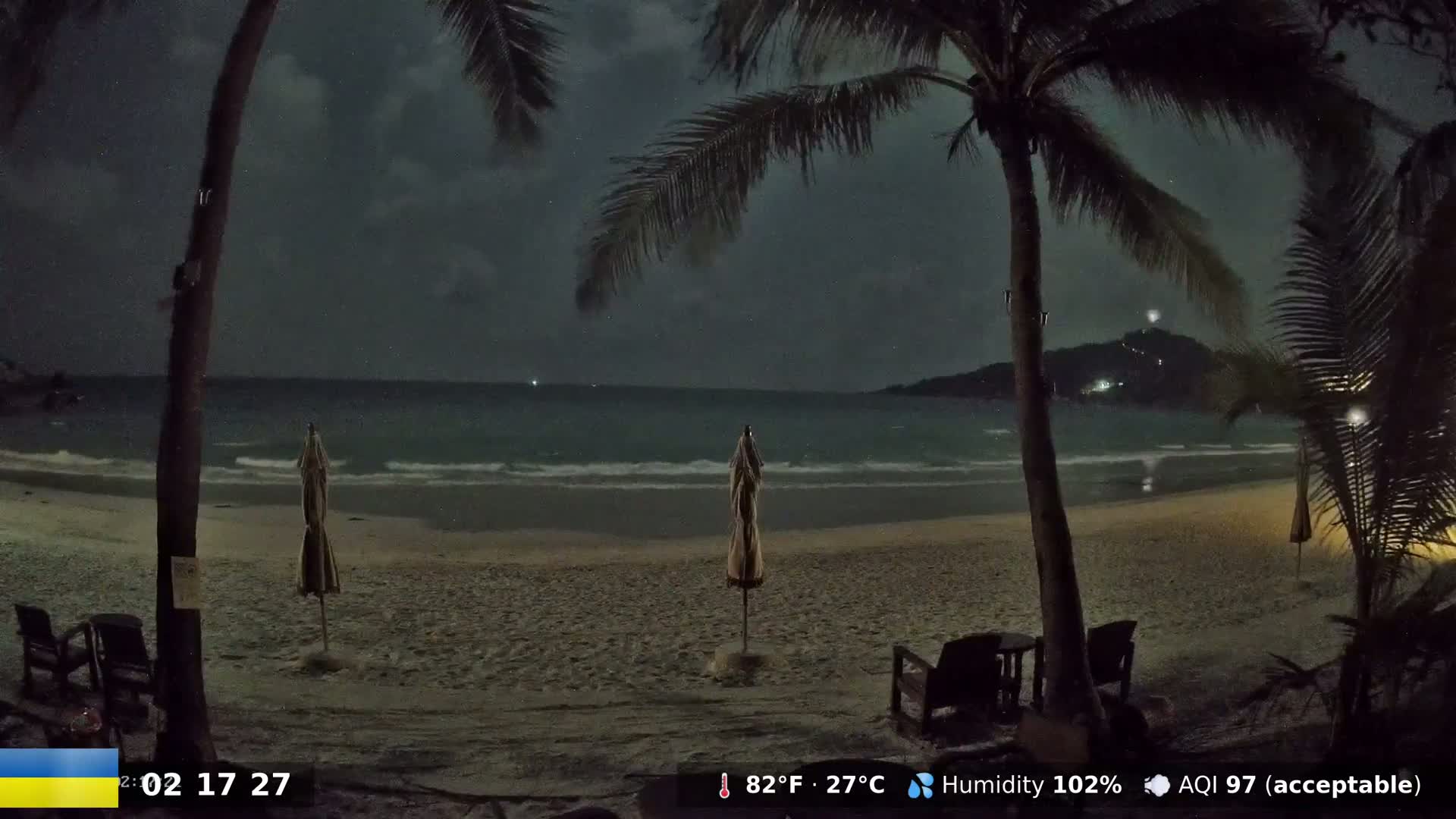 A nighttime beach scene shows a sandy shore with a calm sea under a cloudy sky, palm trees framing the view, and beach umbrellas and chairs on the sand.