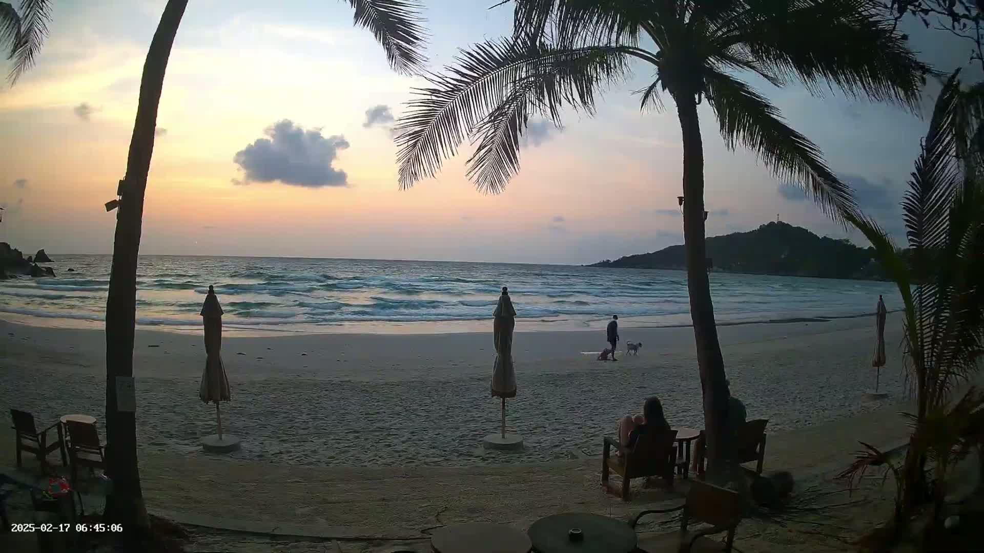 A sandy beach at sunset with gentle waves, palm trees, and people relaxing near beach umbrellas.