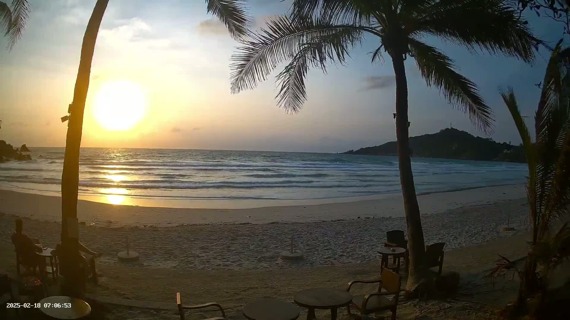 A sandy beach at sunset with gentle waves, palm trees, and outdoor seating.