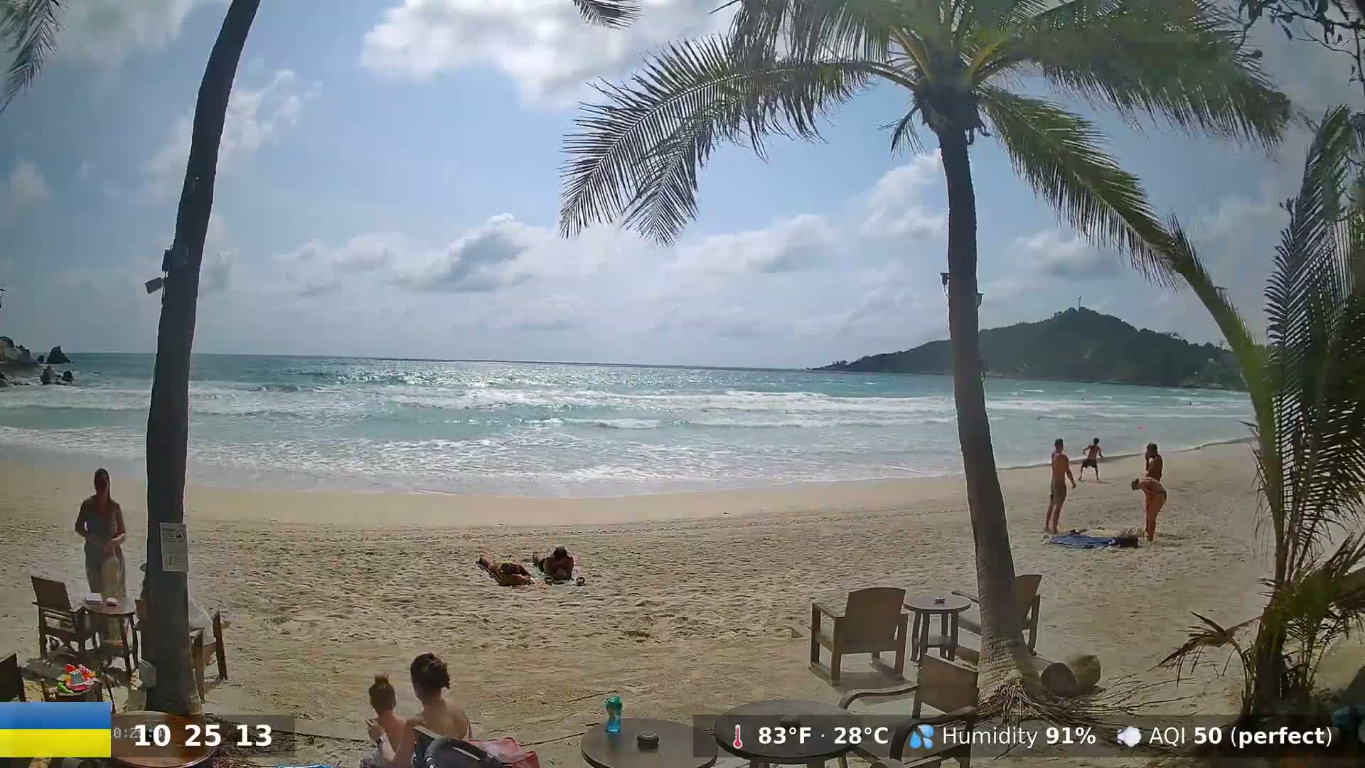 A sunny beach scene shows a sandy shore with calm waves lapping the sand, palm trees, and several people relaxing and swimming in the ocean.