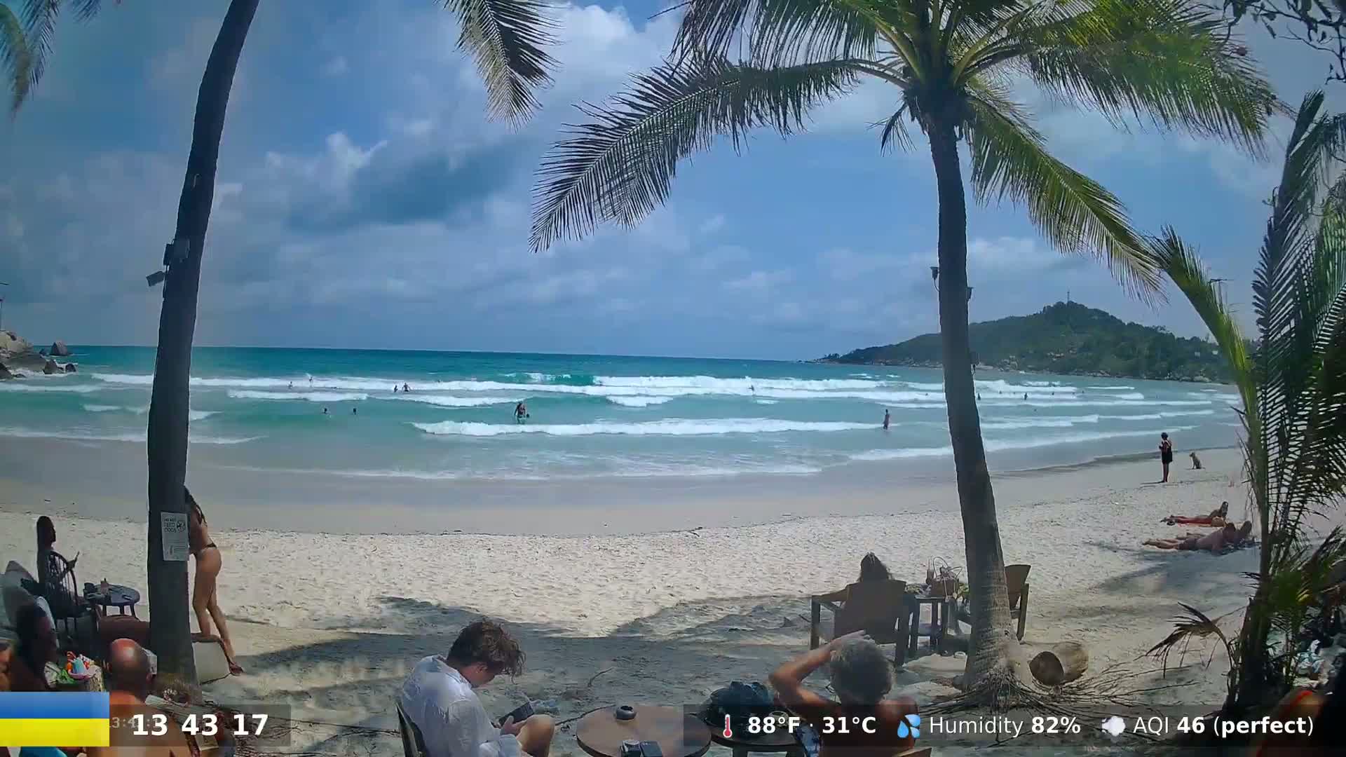 A sandy beach with gentle waves, palm trees, and several people relaxing or swimming under a partly cloudy sky.