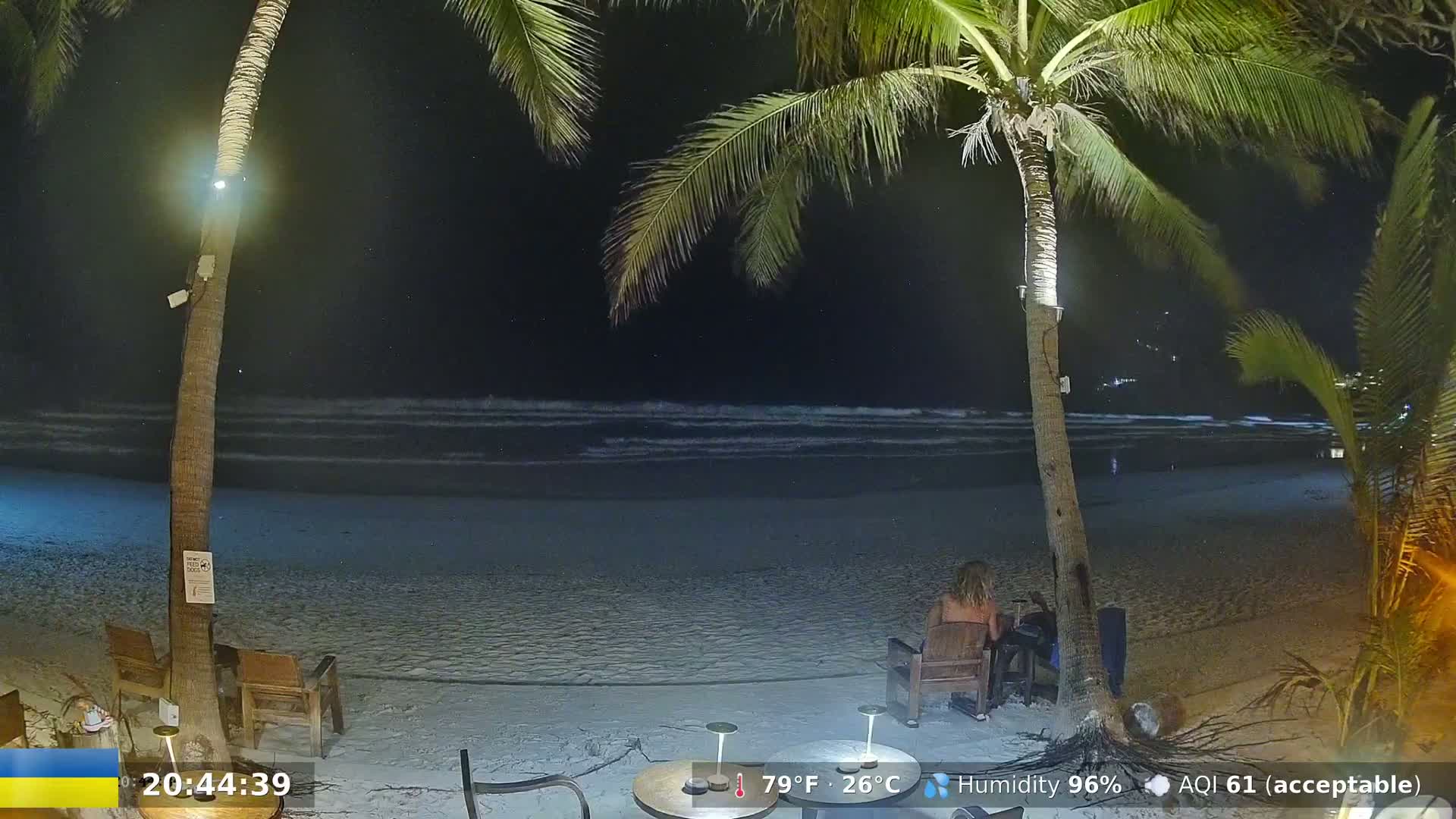A nighttime beach scene shows calm waves lapping a sandy shore under a partly cloudy sky, with two palm trees and people sitting at outdoor tables.