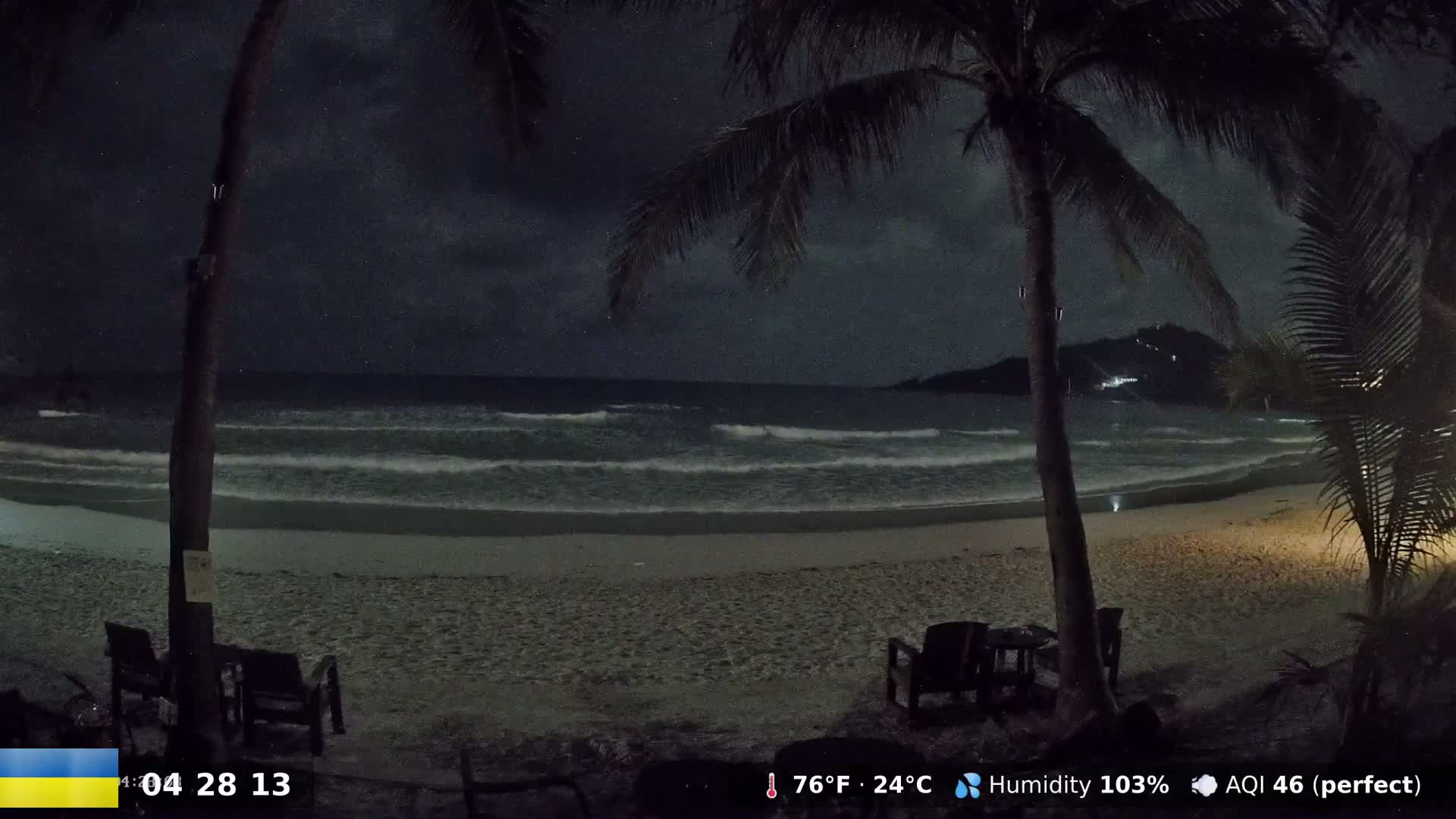 A nighttime beach scene under a cloudy sky shows gently lapping waves on a sandy shore with palm trees and empty beach chairs.
