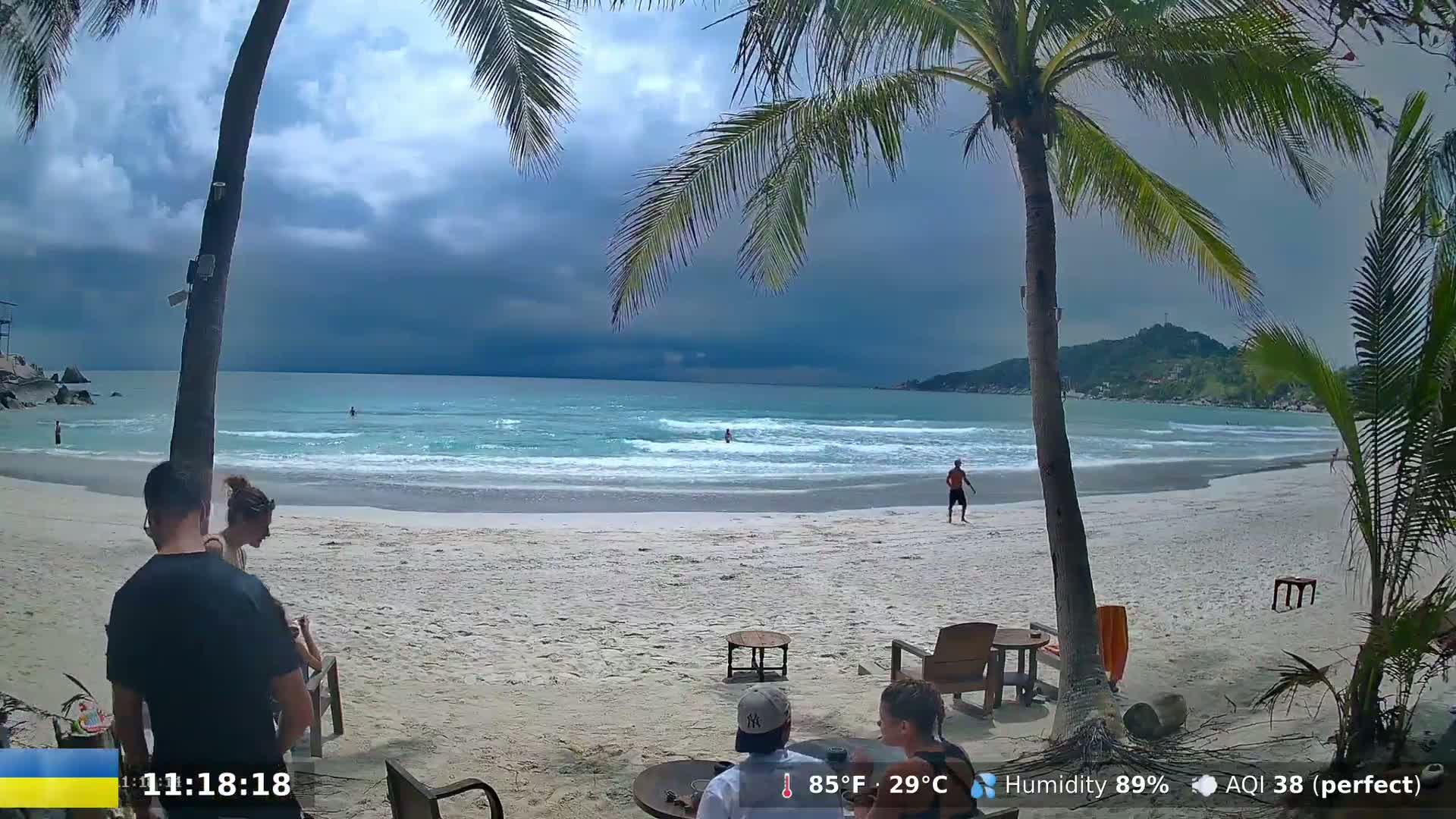 A tropical beach scene under a partly cloudy sky shows people relaxing on the sand near palm trees and beach furniture.