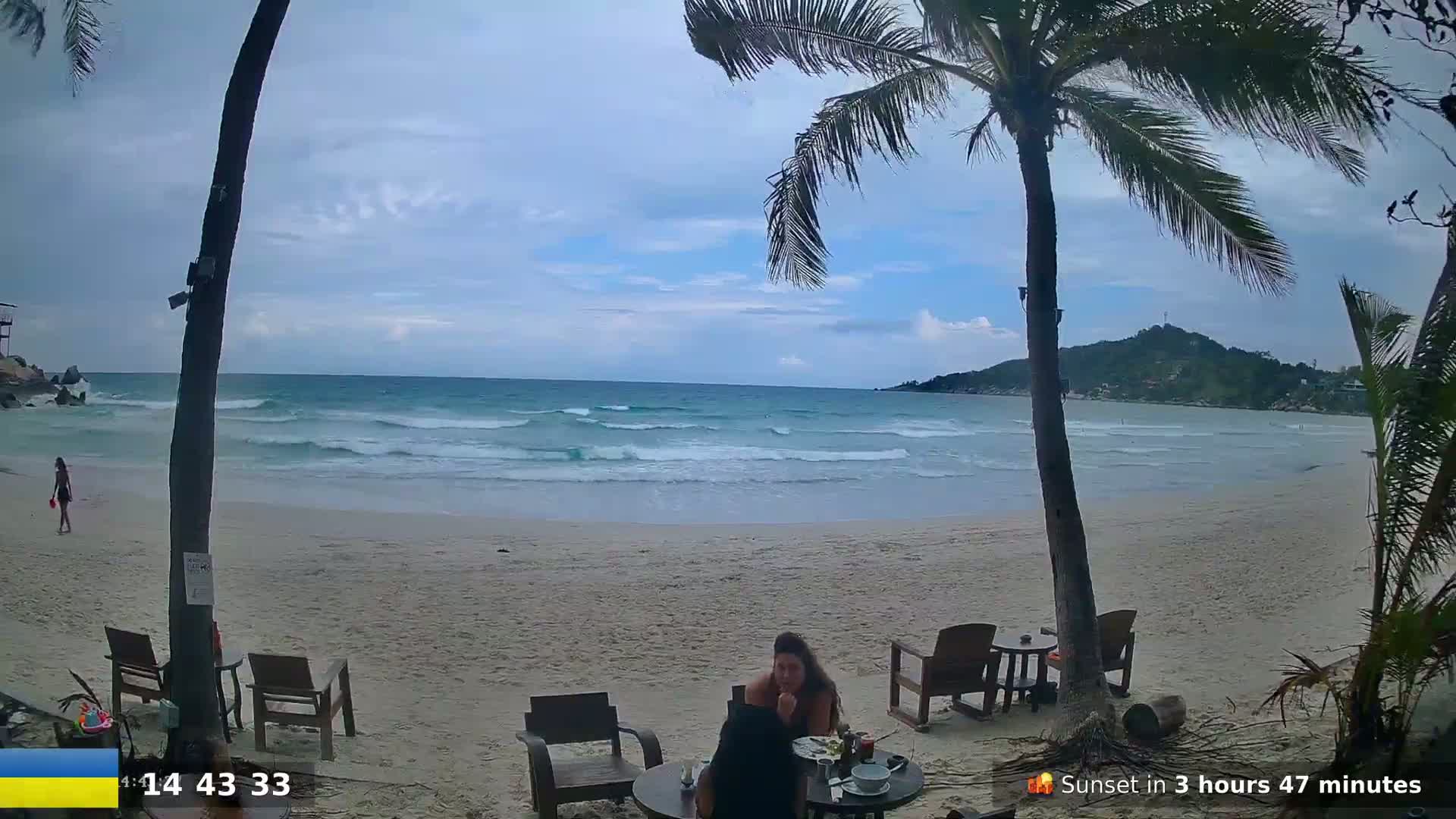 A sandy beach with gentle waves, palm trees, and a couple sitting at an outdoor table under an overcast sky.