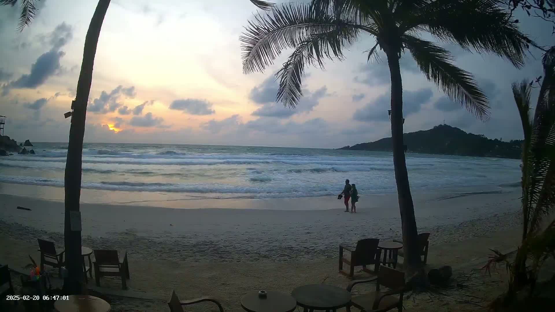 Two silhouetted figures stand on a sandy beach at sunset as small waves lap the shore, framed by palm trees and a distant island.