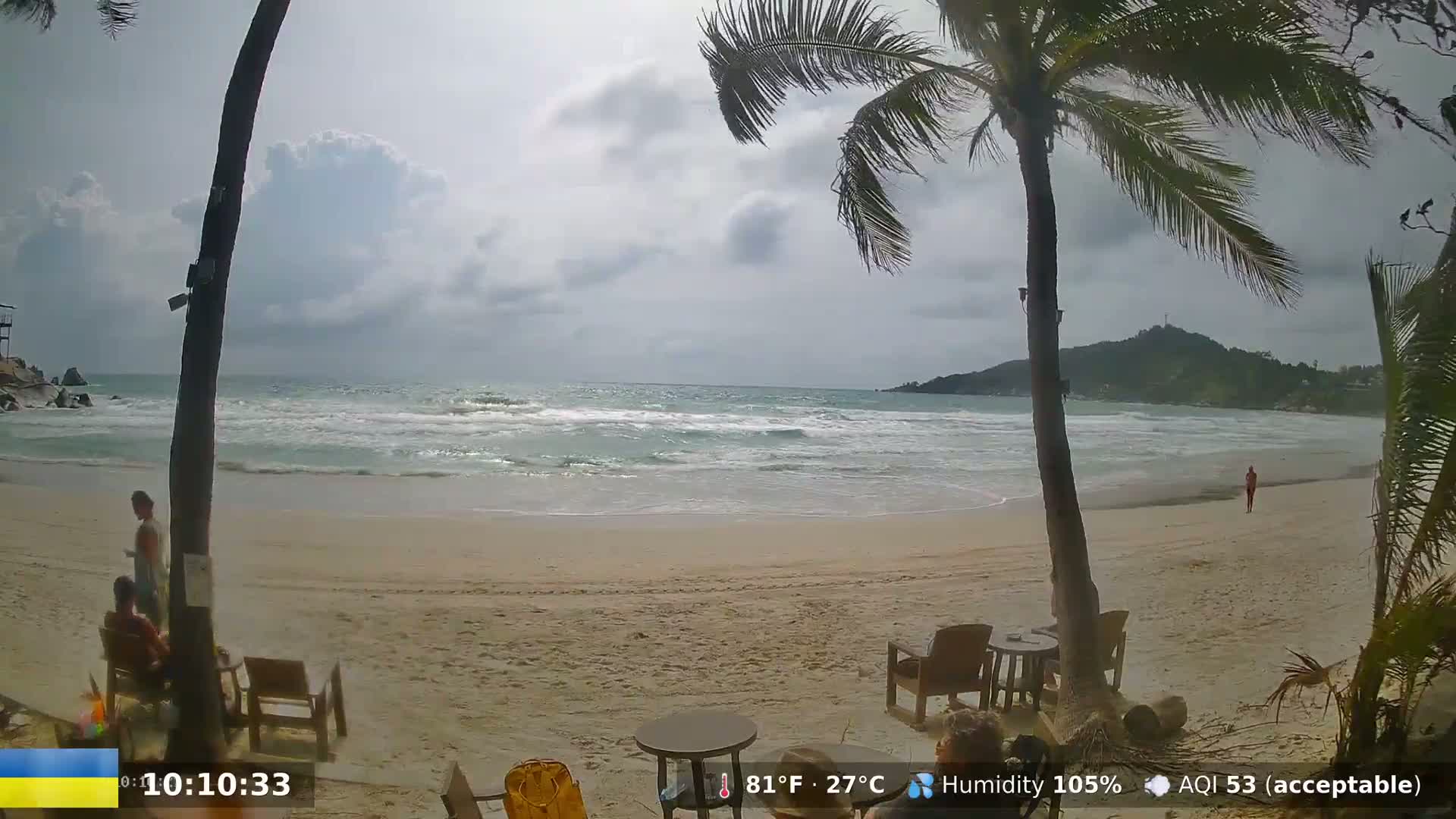 A sandy beach with some people relaxing near palm trees under a partly cloudy sky, with moderately choppy waves in the ocean.