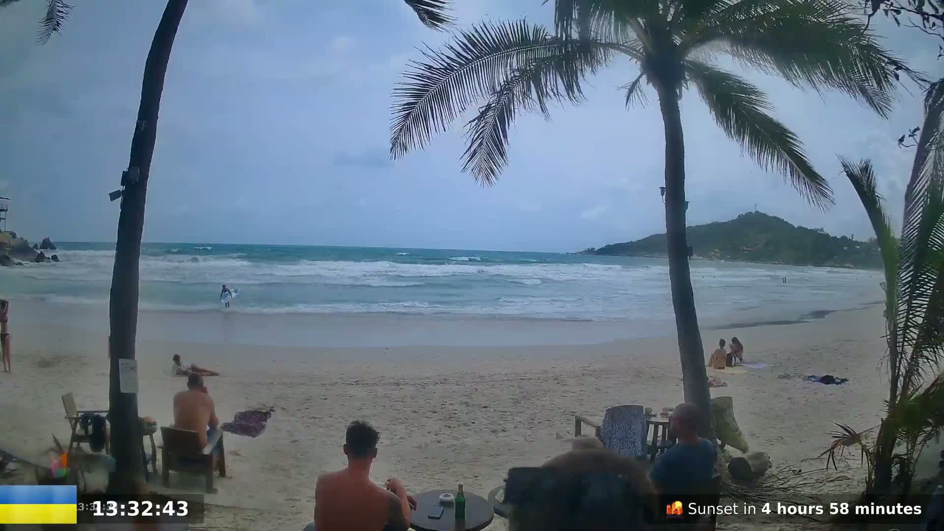A sandy beach with a few people relaxing and one person surfing in moderately wavy ocean water under an overcast sky, partially obscured by palm trees.
