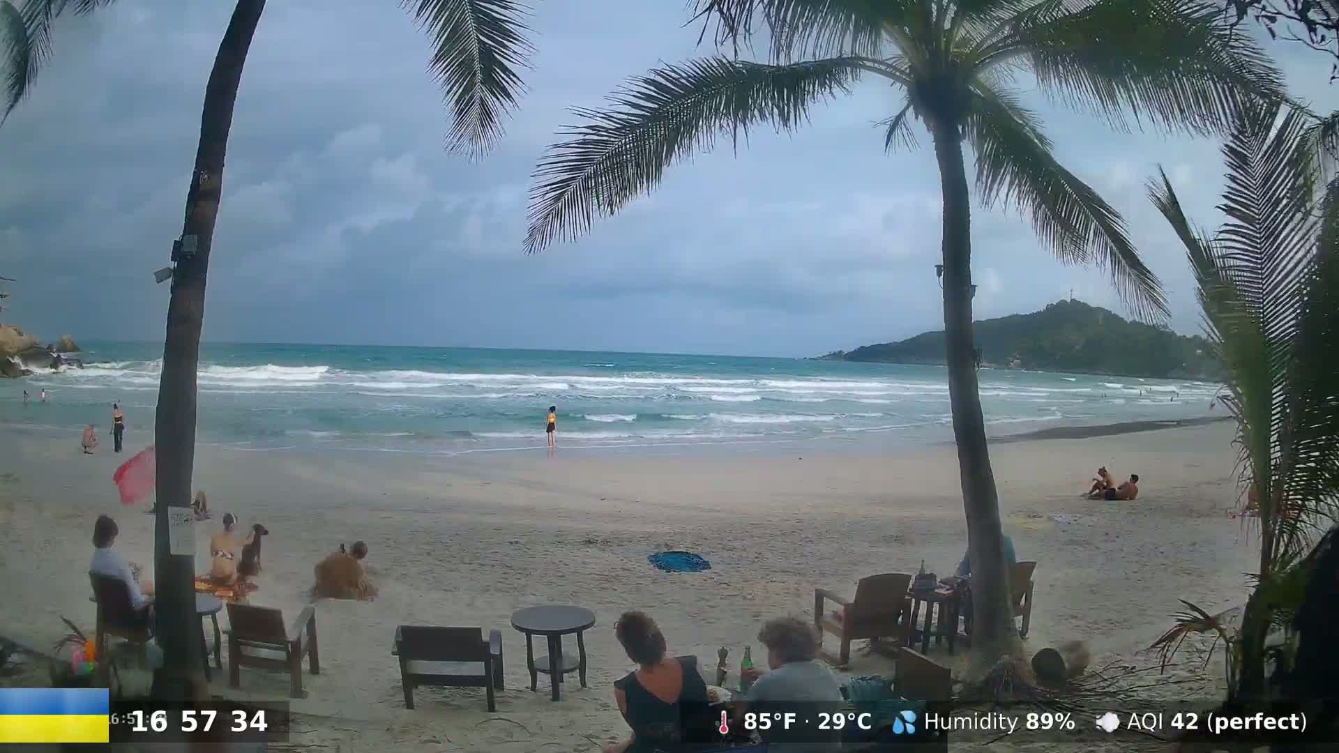 A sandy beach with people relaxing near palm trees under a cloudy sky, with moderate waves in the ocean.