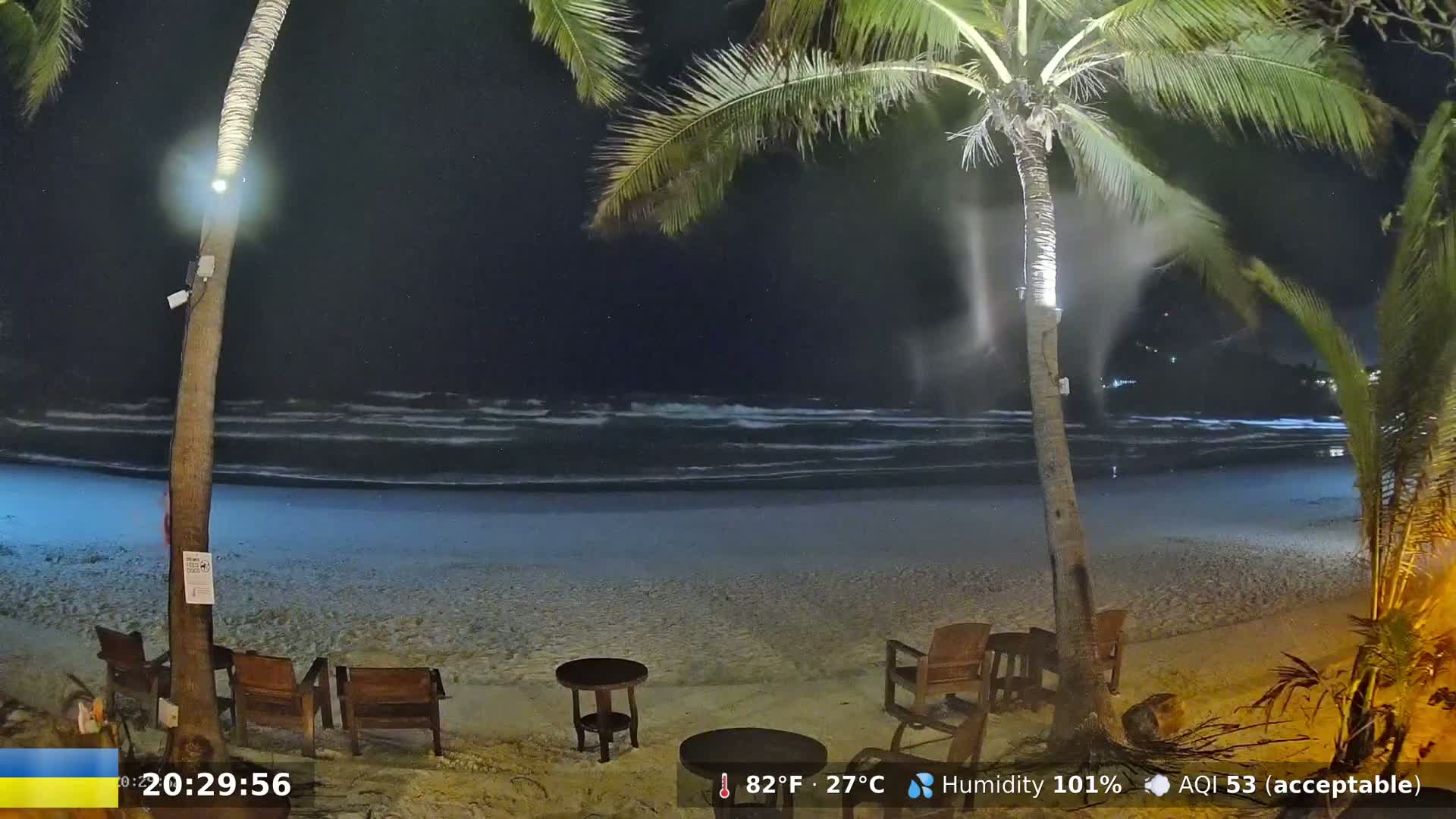 A nighttime beach scene shows a sandy shore with gentle waves, palm trees illuminated by lights, and several empty wooden chairs and a small table.