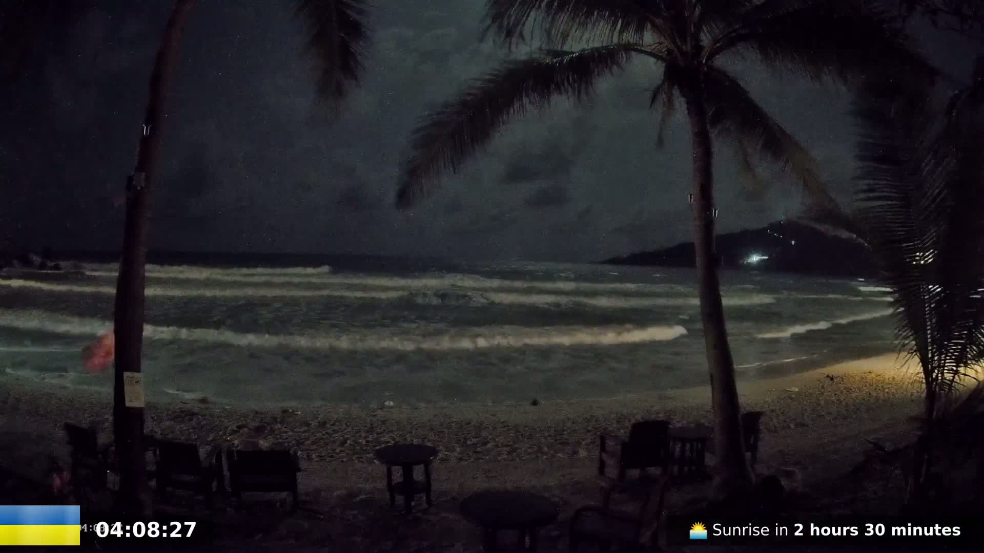 A nighttime beach scene shows moderately sized waves lapping a sandy shore under a dark, cloudy sky, with palm trees and outdoor furniture visible in the foreground.