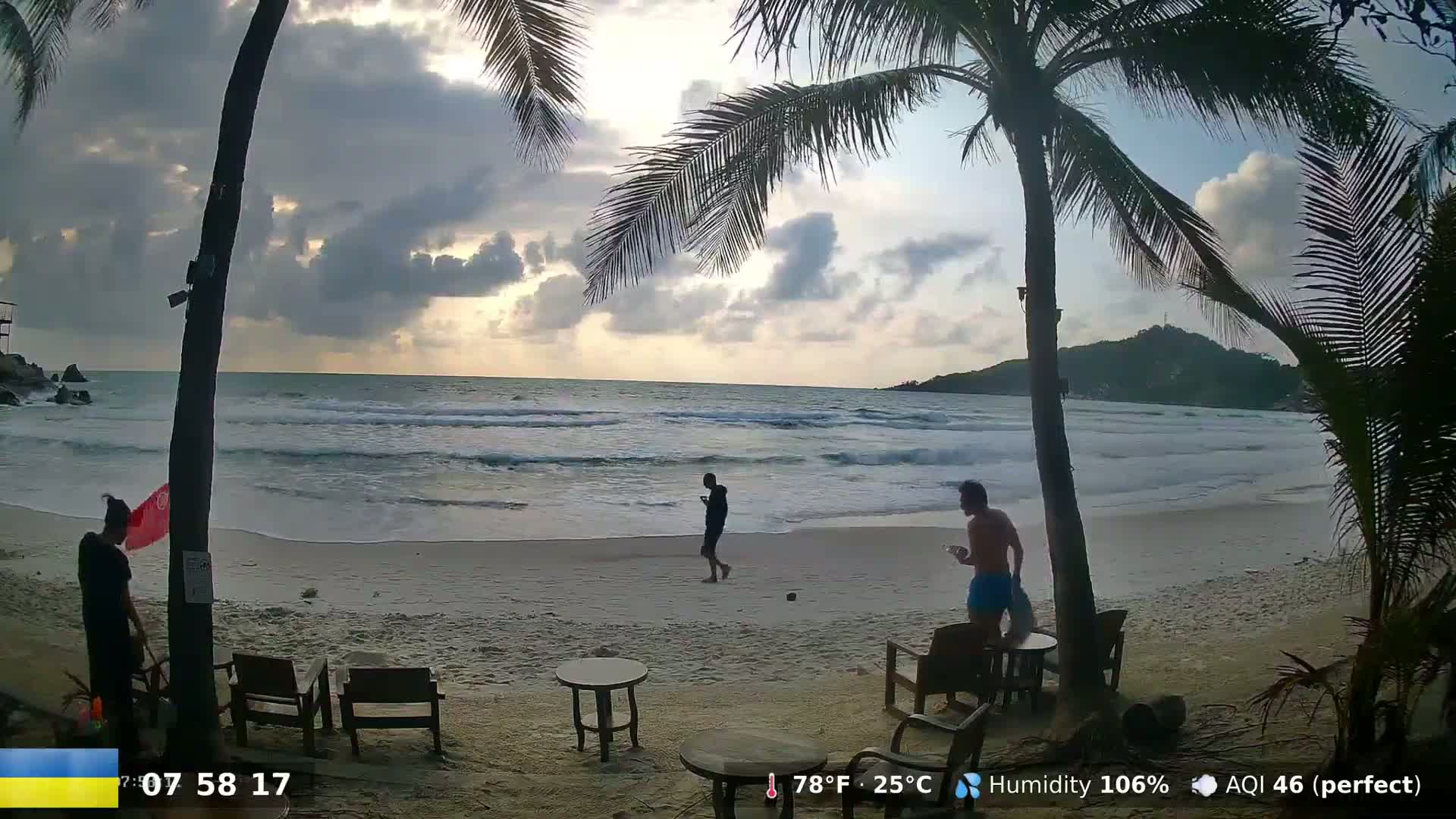 A partly cloudy sunset illuminates a sandy beach with gently rolling waves, palm trees, and several people near beach chairs and tables.