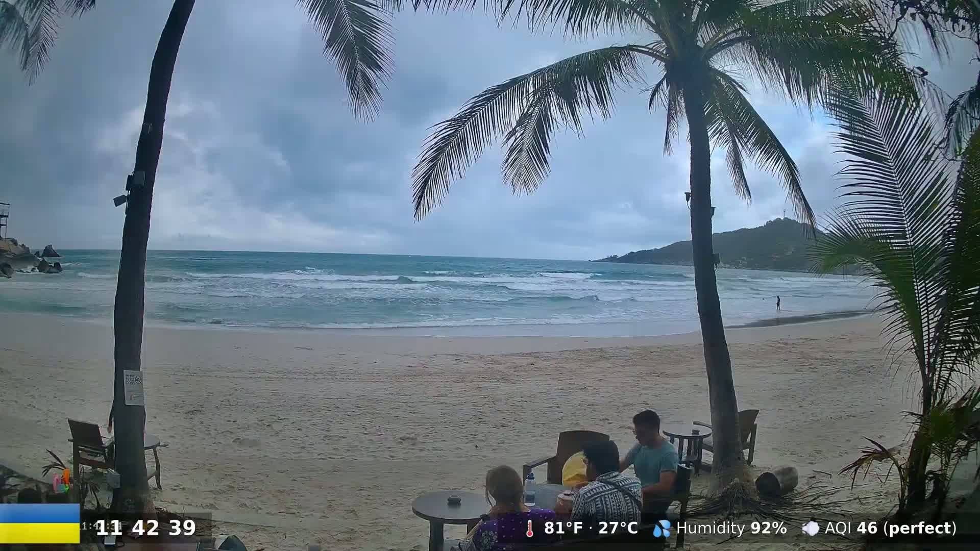 A sandy beach with moderate waves under an overcast sky, palm trees, and several people sitting at an outdoor table.