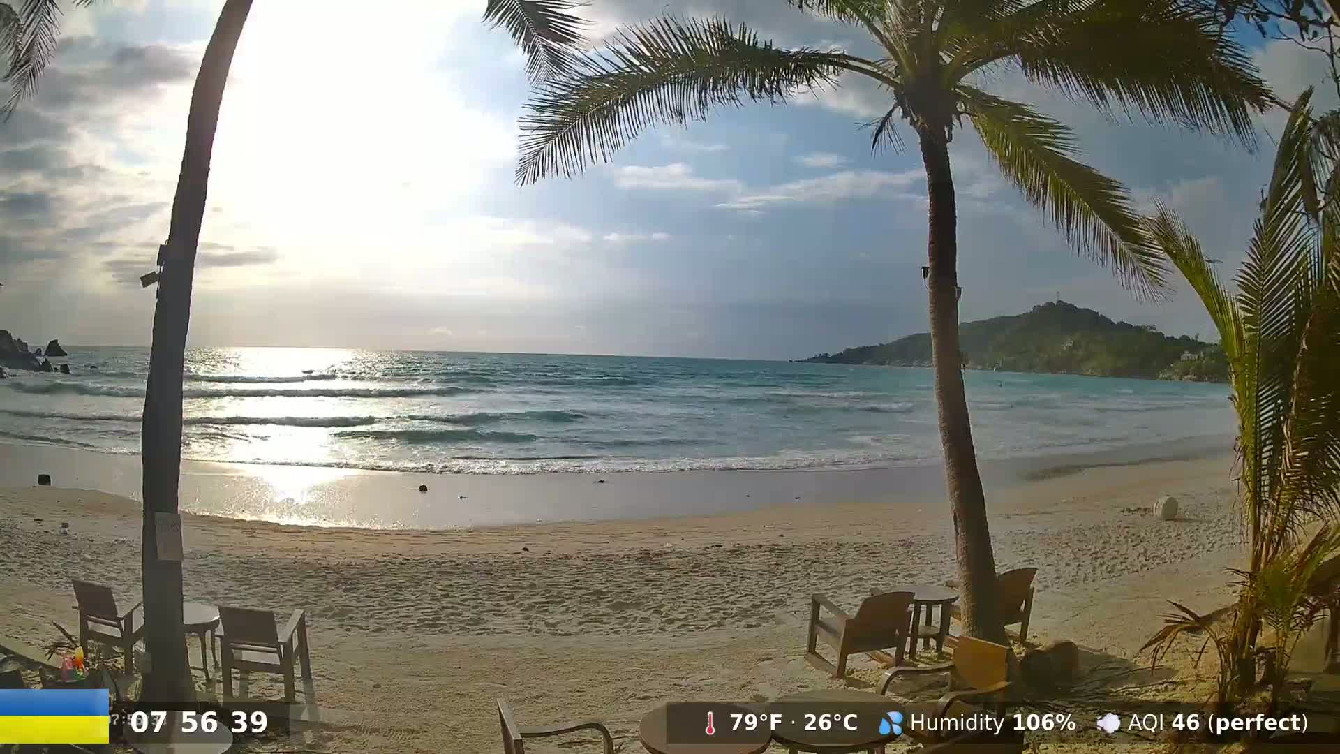 A partly sunny beach scene shows a sandy shore with gentle waves, palm trees, and outdoor furniture under a mostly cloudy sky.