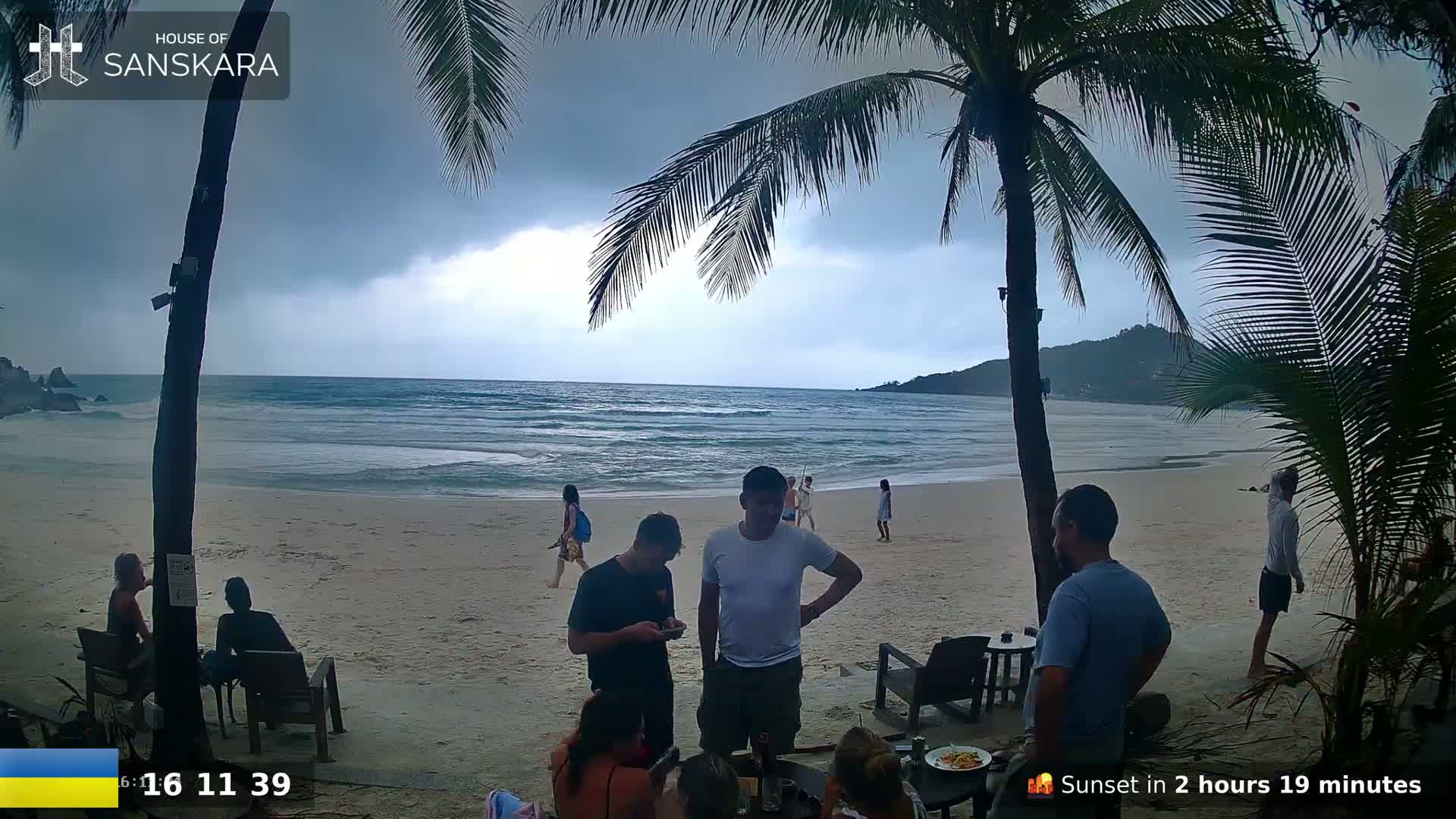 A sandy beach with calm ocean waves under an overcast sky, palm trees, and several people relaxing near beach furniture.