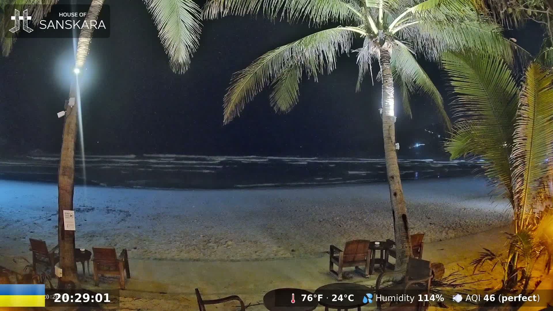 A nighttime beach scene shows gently lapping waves under a clear, starry sky, with palm trees and outdoor seating visible near the shore.