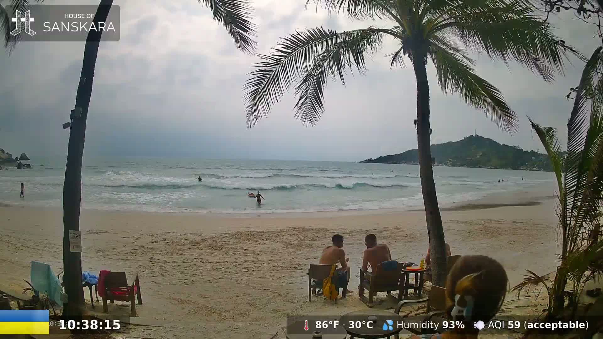 A sandy beach with moderate waves under an overcast sky, shows people swimming in the ocean and others relaxing at outdoor beach furniture under palm trees.