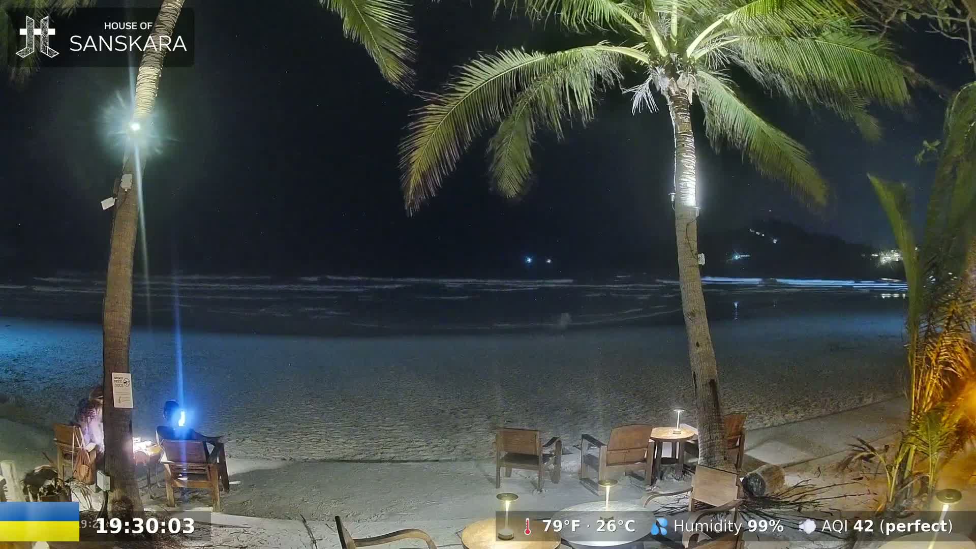 A nighttime beach scene shows gently lapping waves, palm trees, outdoor furniture, and two people sitting at a table under the starry sky.
