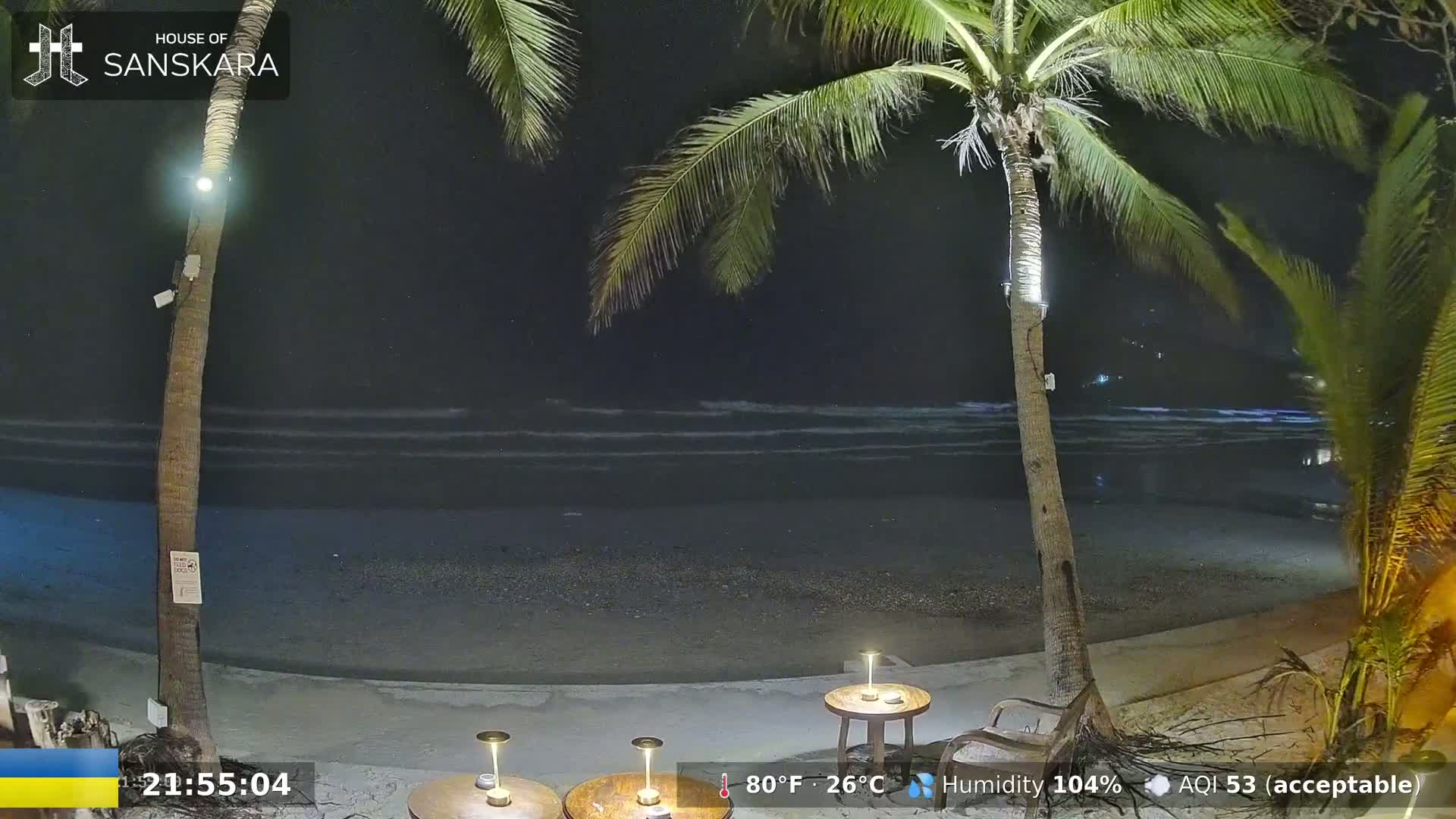 At night, a dimly lit beach scene shows a dark ocean with gentle waves, a sandy shore, two palm trees, and small tables with lamps.