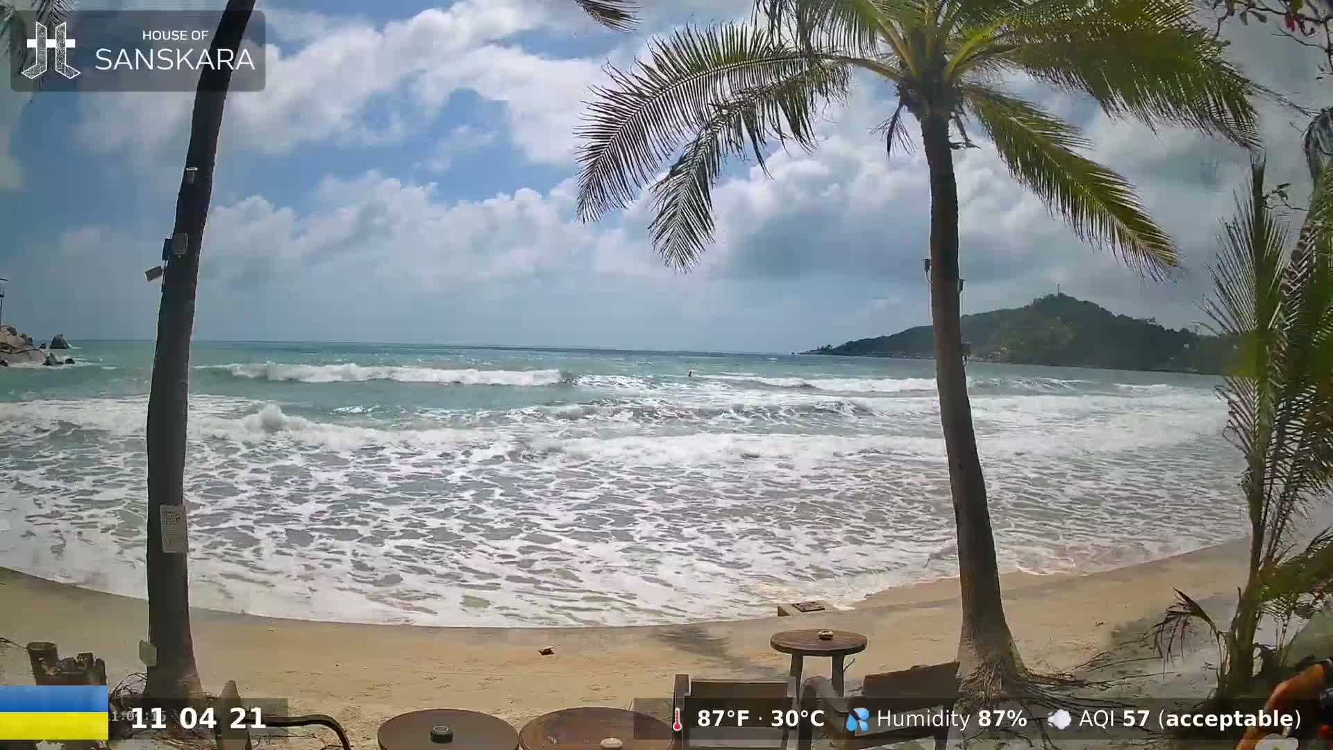 A sandy beach with moderate waves under a partly cloudy sky, palm trees are in the foreground.