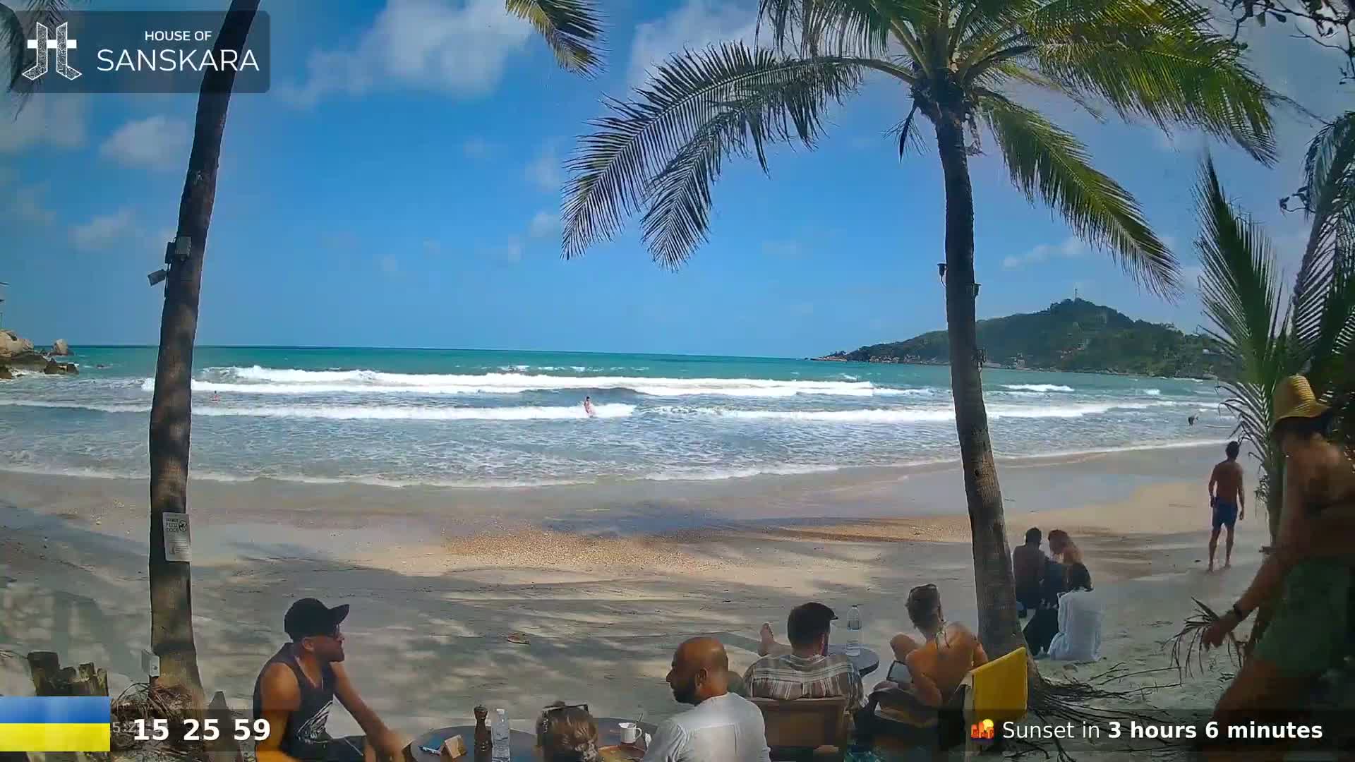 A sandy beach with moderate waves under a bright, sunny sky, palm trees, and several people relaxing on the sand and in the water.