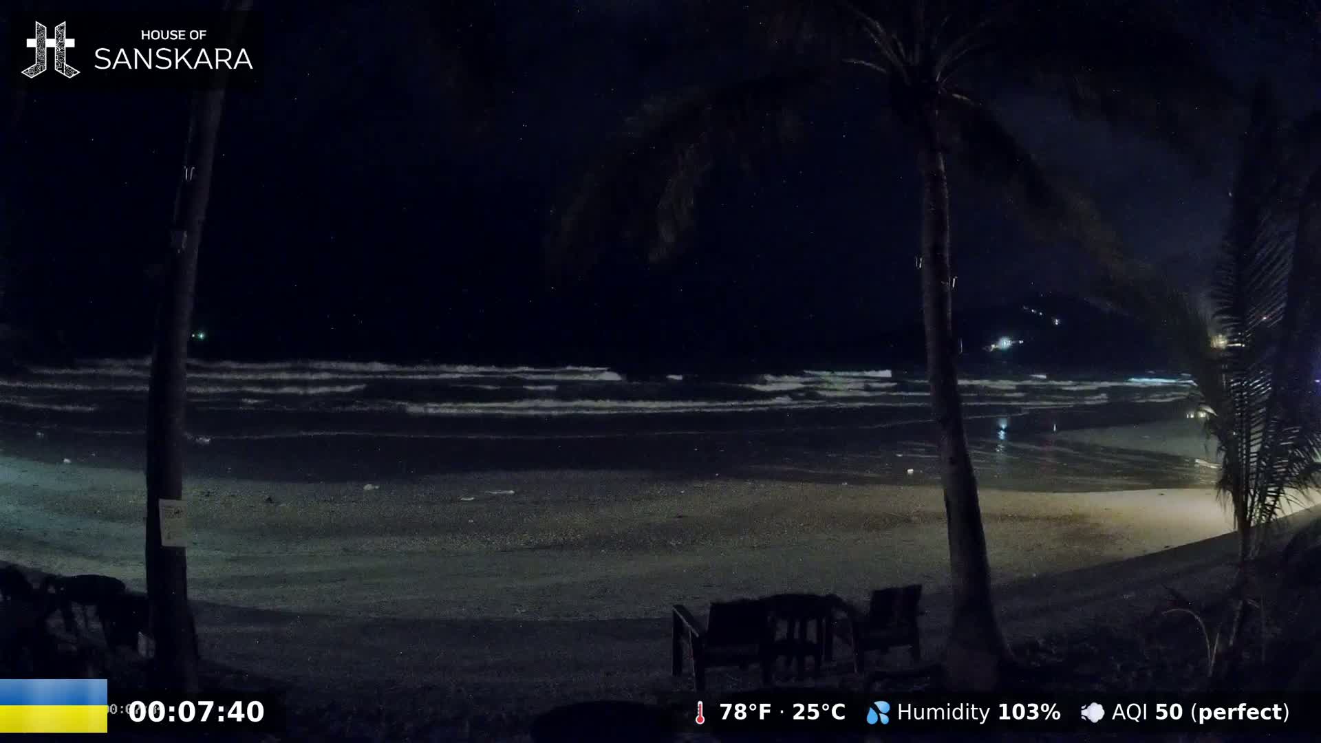 A nighttime beach scene shows gently lapping waves under a dark, starry sky, with palm trees in the foreground and empty chairs on the sand.