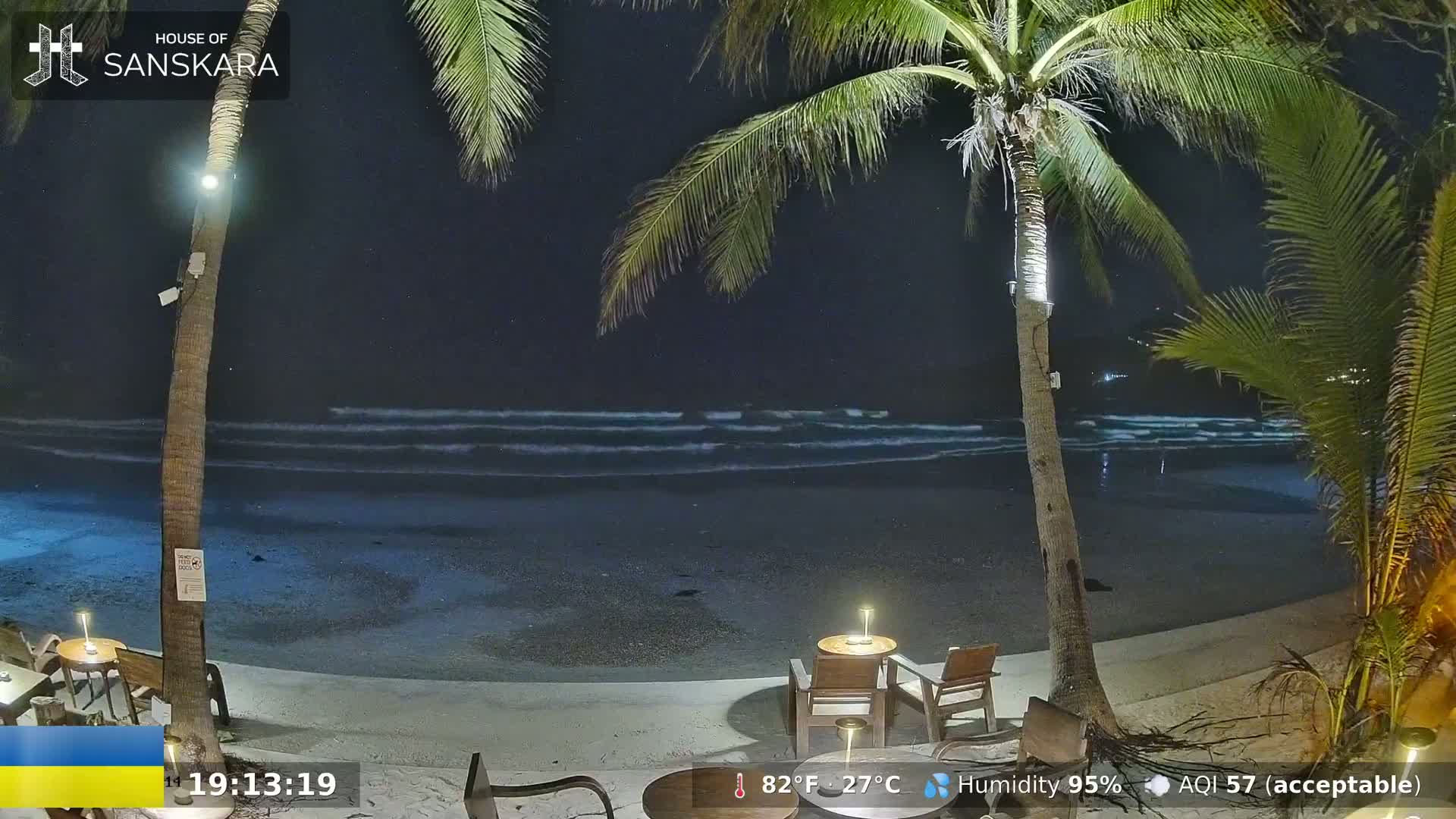 A nighttime beach scene shows gently lapping waves under a dark sky, with palm trees and outdoor seating visible in the foreground.