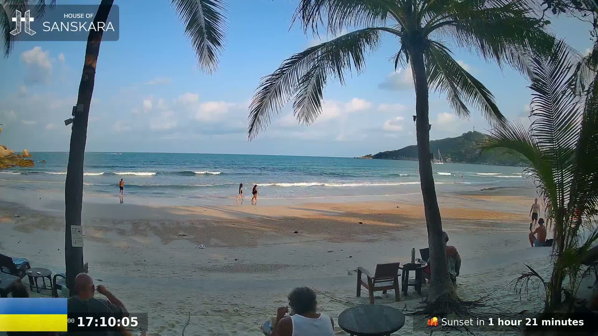 A sandy beach with calm ocean waves, palm trees, and several people are visible under a partly cloudy sky.