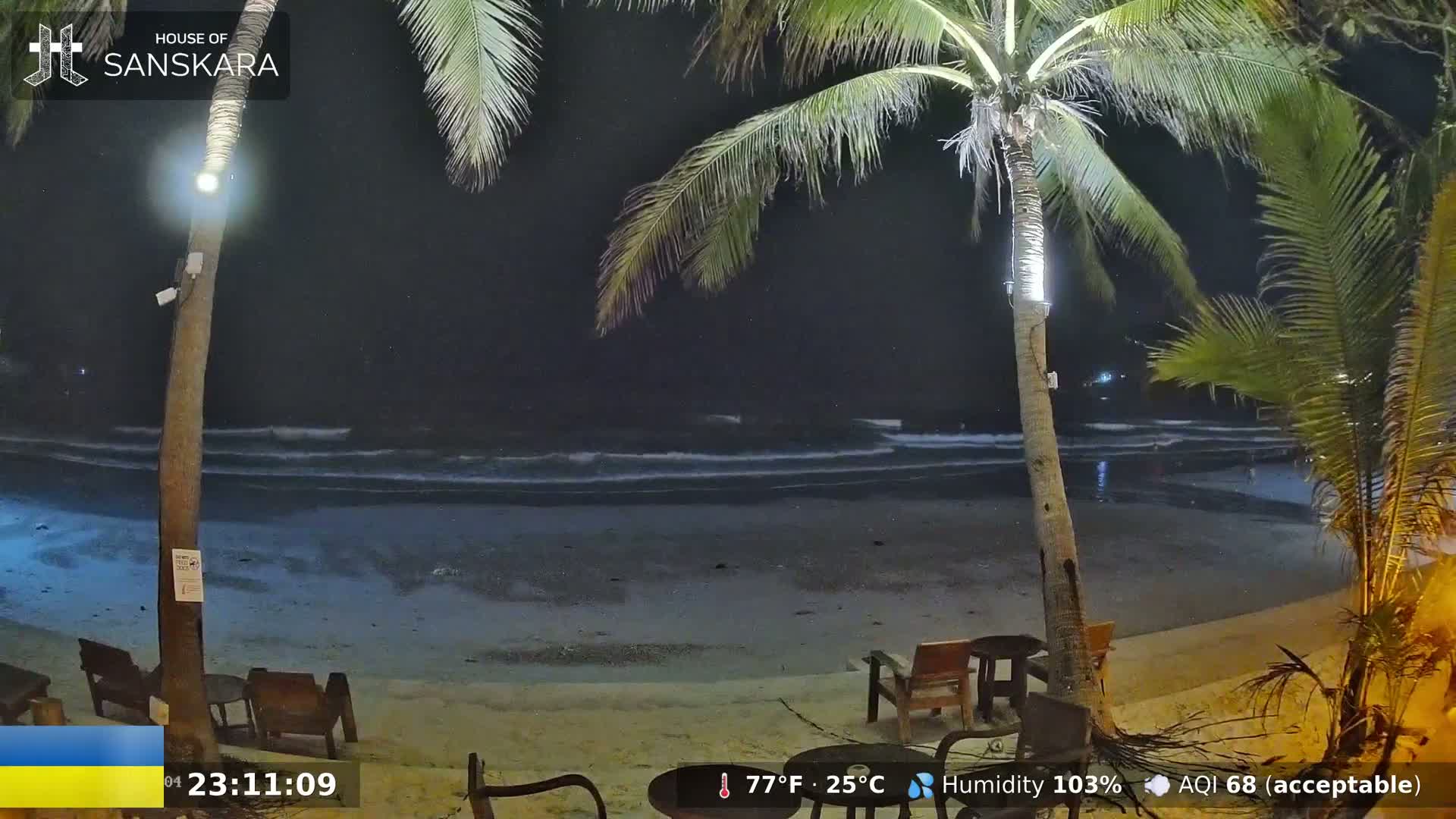 A nighttime beach scene shows gently lapping waves under a dark sky, with palm trees and outdoor seating illuminated on the sand.